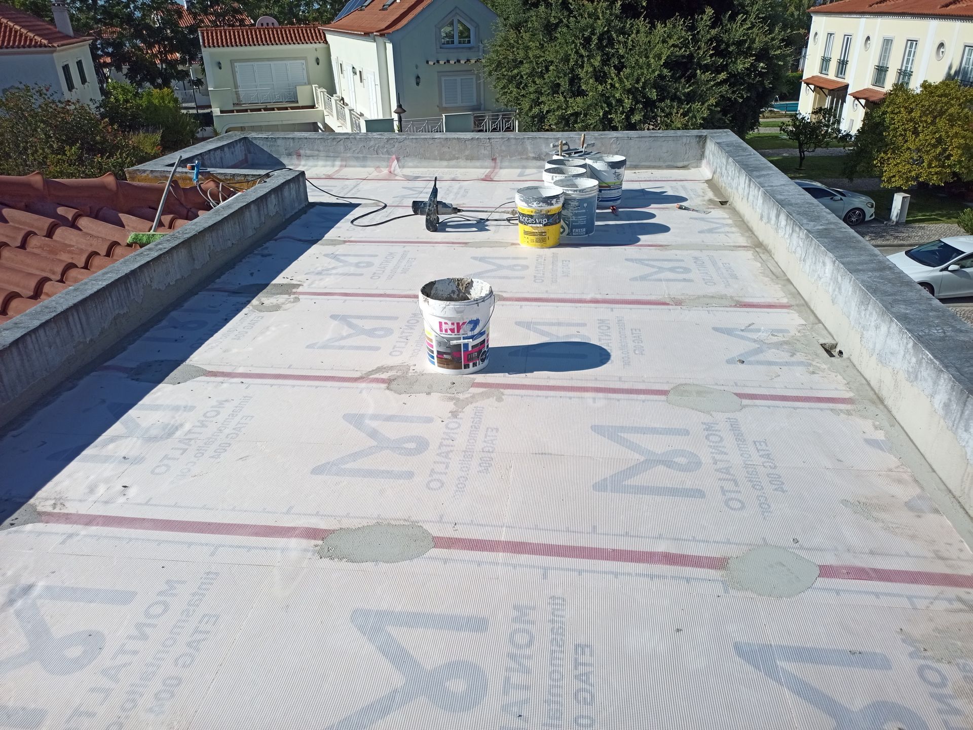 Telhado plano com materiais de construção e borda de concreto. Superfície branca com linhas vermelhas, balde e equipamentos.