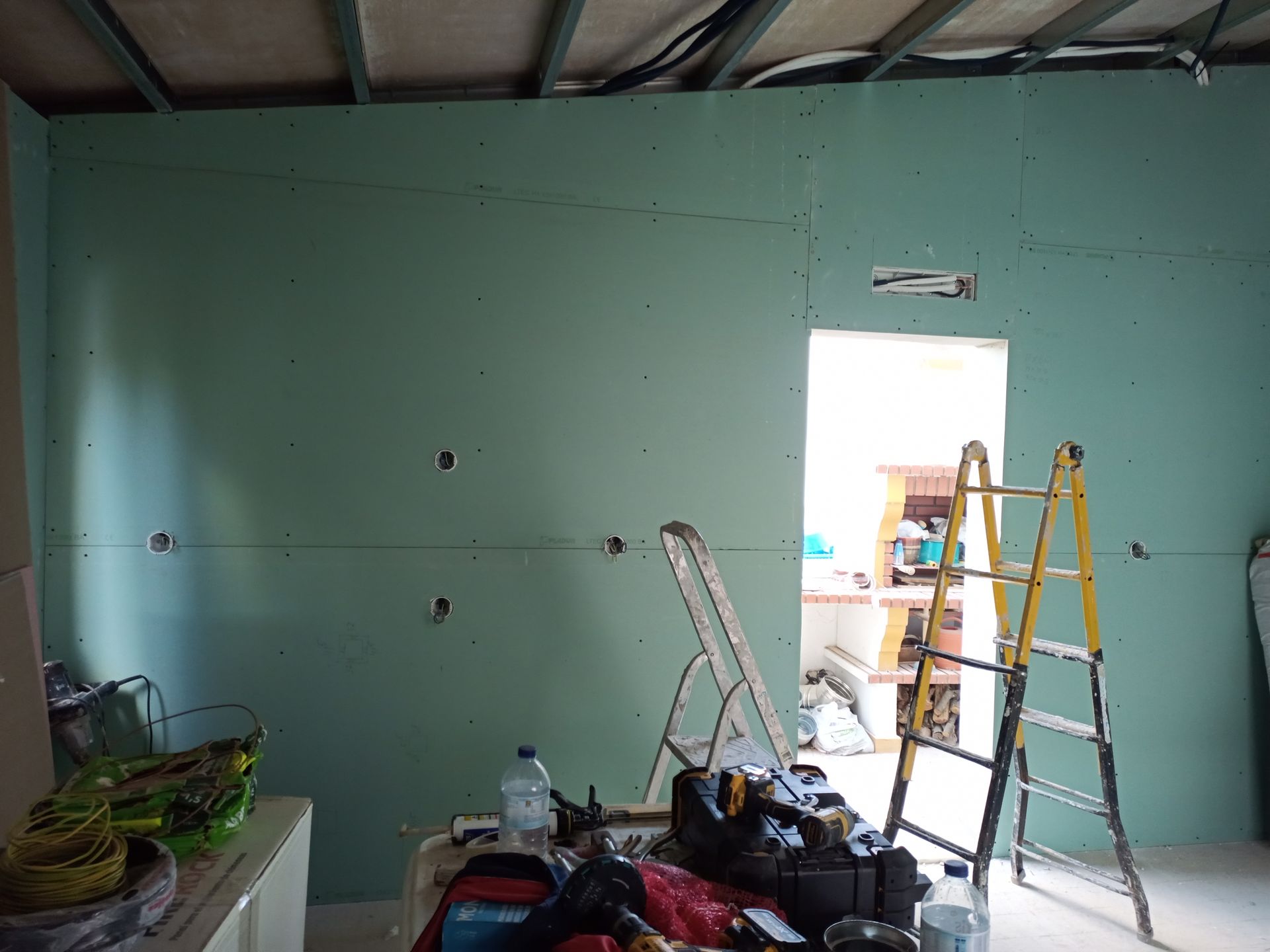 Interior com paredes de gesso verde, uma porta aberta, escadas e ferramentas, em uma sala em construção.