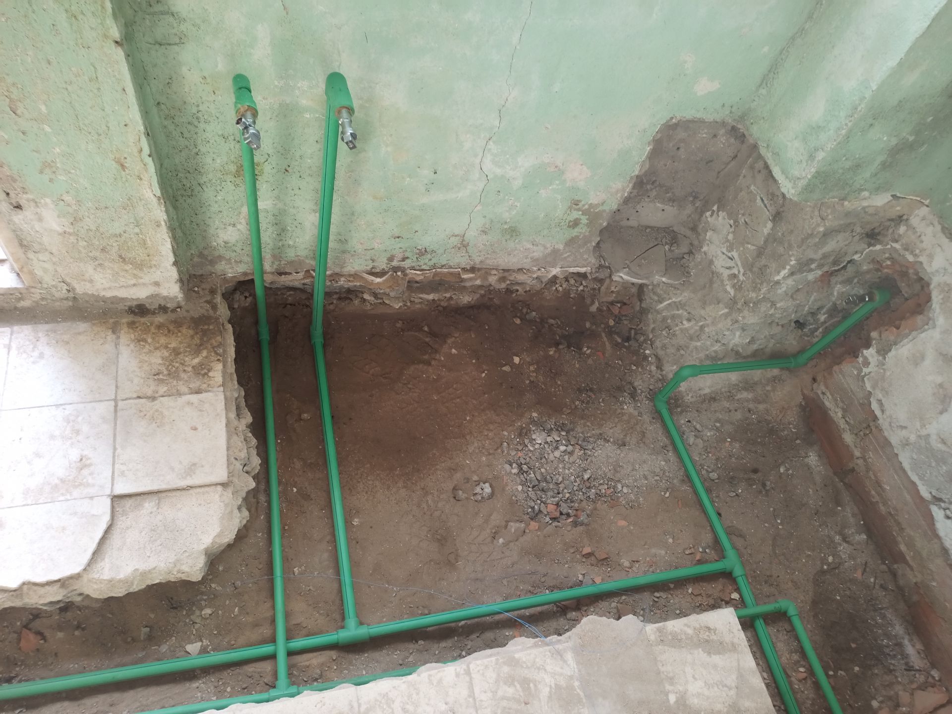 Tubulações verdes expostas em um canteiro de obras; piso de terra, área revestida com azulejos e paredes danificadas.