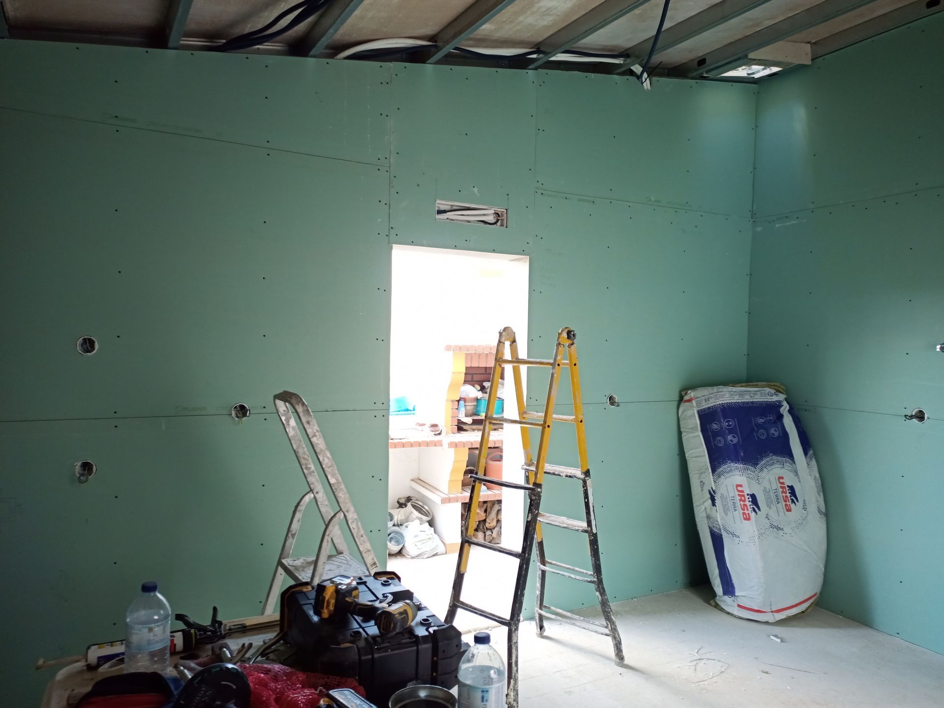 Quarto interno em construção, com drywall verde, porta aberta, escada e ferramentas.