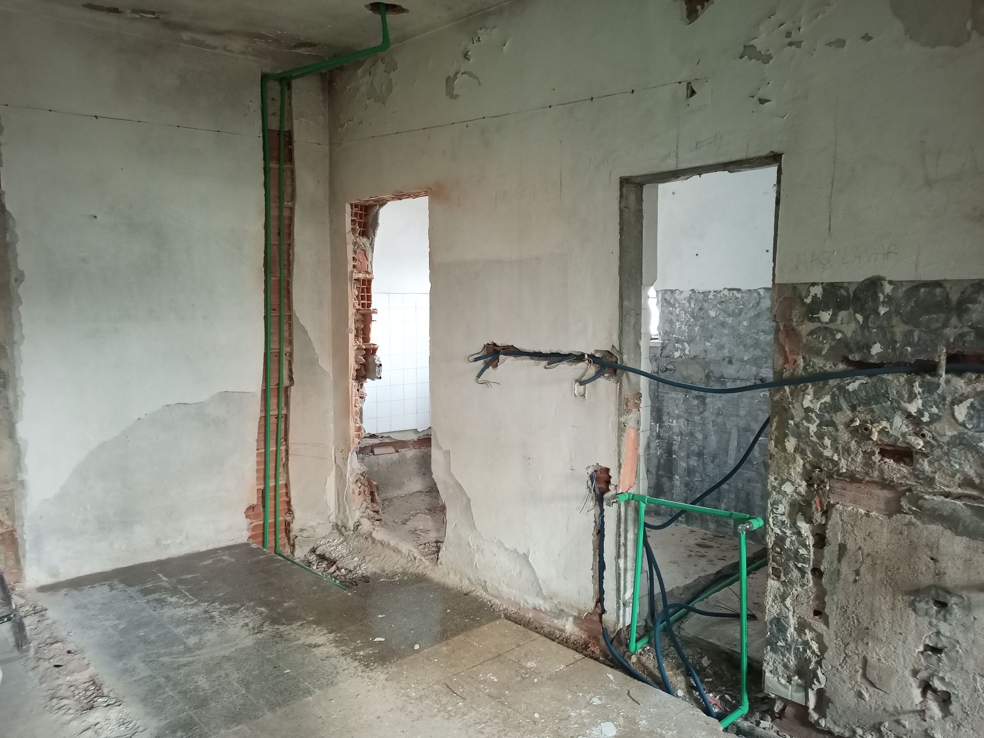 Quarto interior em construção com tubulações de encanamento e portas expostas. Piso de concreto e paredes danificadas.