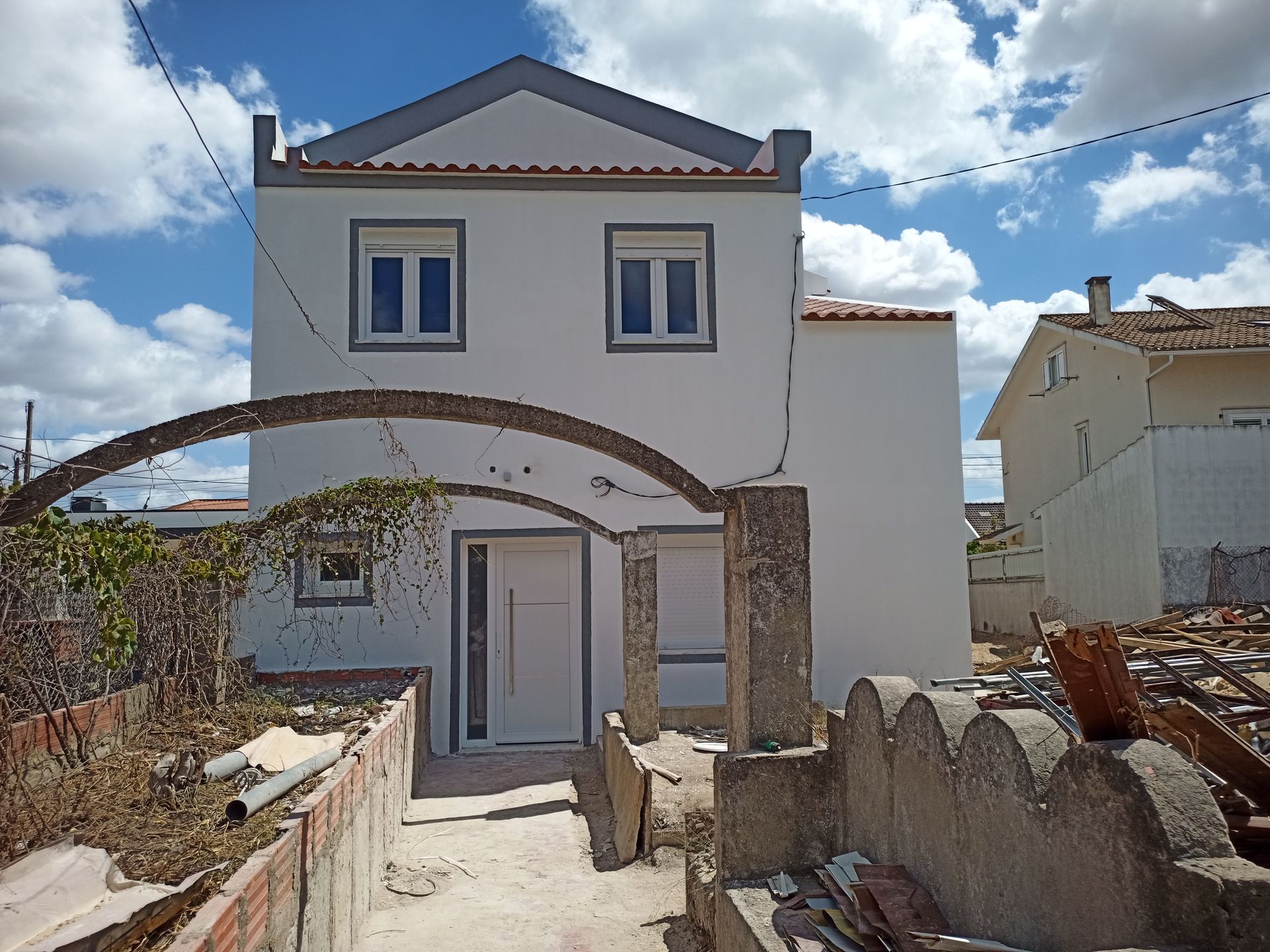 Casa branca de dois andares com telhado cinza, entrada em arco e detritos em primeiro plano sob um céu nublado.
