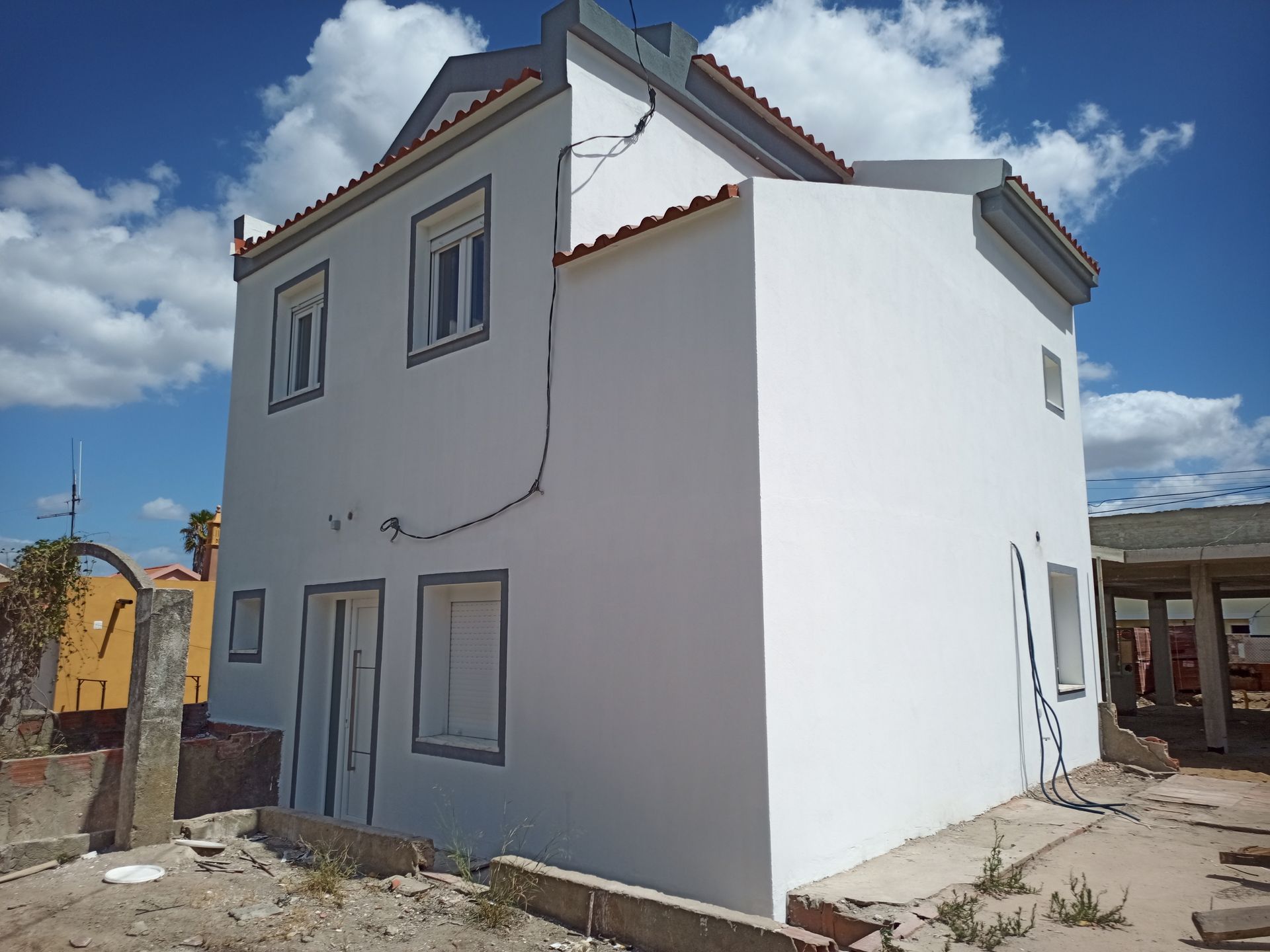 Casa branca de dois andares com telhado de telha vermelha contra um céu azul. Entulho de construção em primeiro plano.