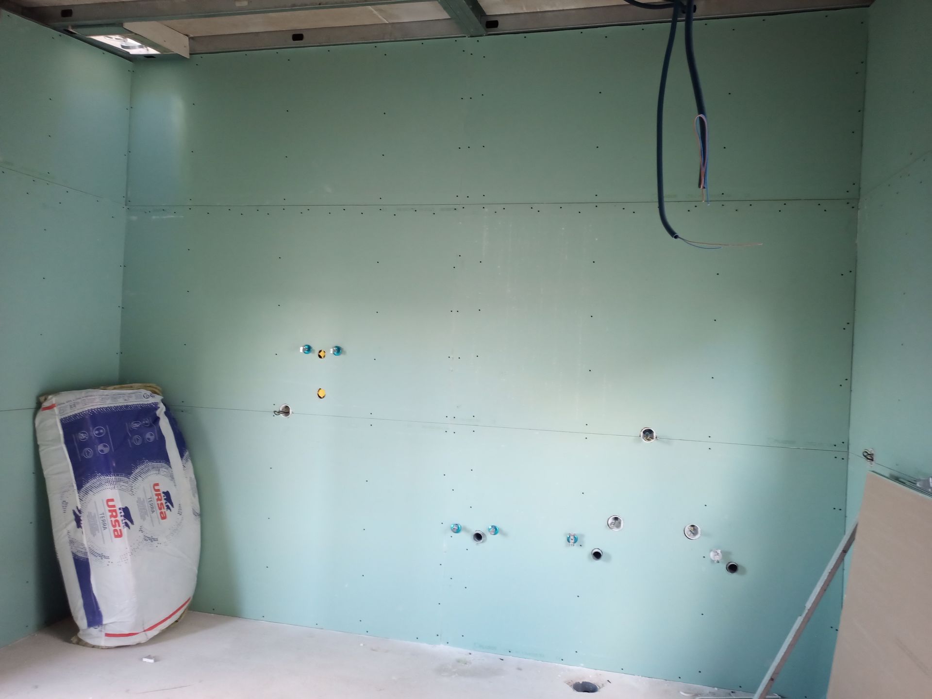 Interior de um cômodo em construção com paredes de gesso acartonado verde. Uma sacola de materiais de construção está em um canto, com fiação exposta na parede.