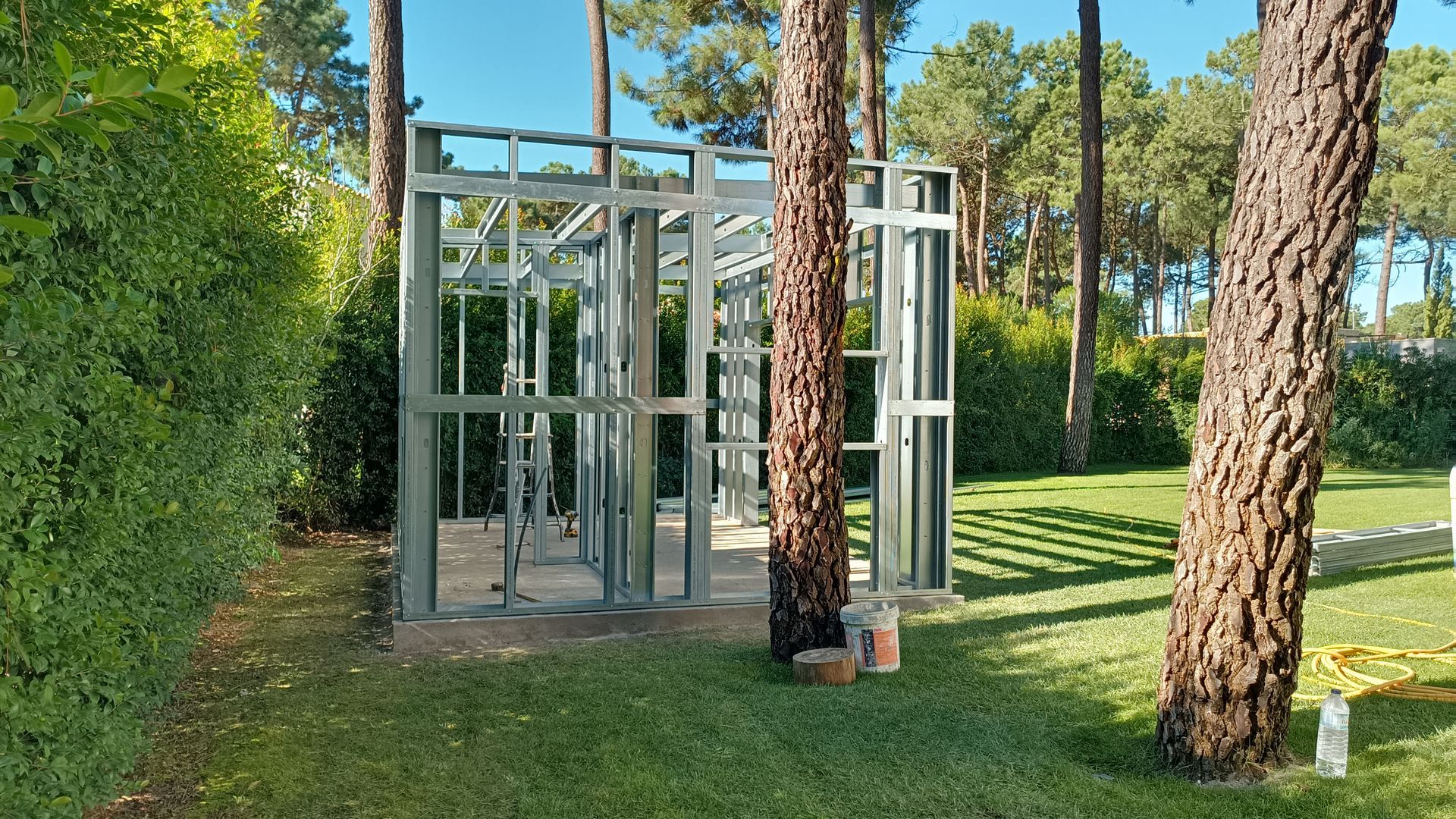 Estrutura metálica de um edifício em construção, situada num pátio gramado com árvores.