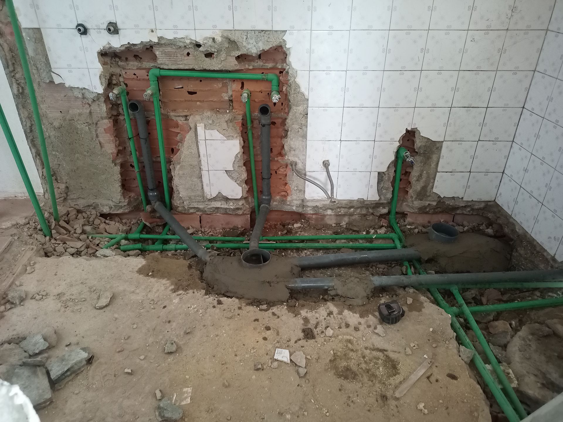 Tubos verdes e cinzentos expostos numa abertura na parede, provavelmente para trabalhos de canalização numa divisão com paredes revestidas a azulejos e piso de concreto.