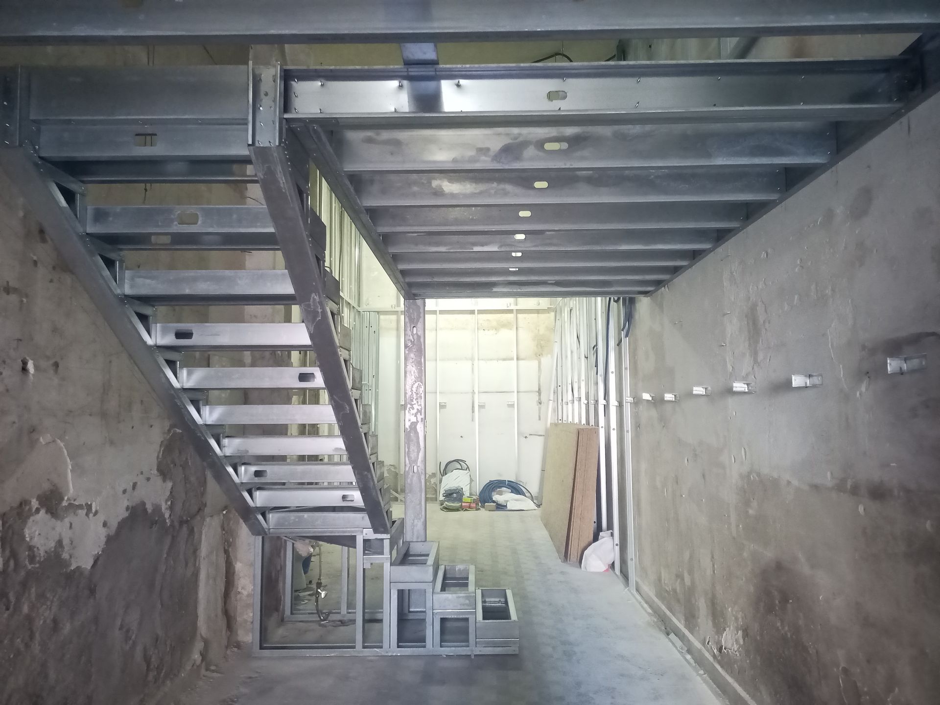 Escada metálica em construção dentro de um cômodo. Paredes e suportes aparentes, com materiais ao fundo.