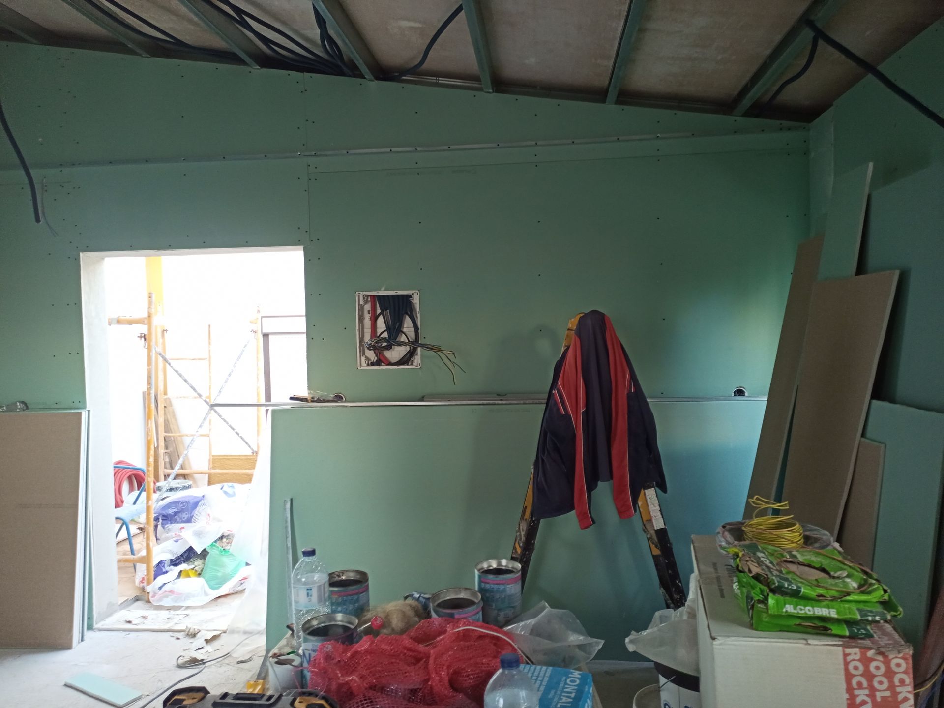 Interior de uma sala em construção, com paredes verdes, porta aberta e ferramentas/materiais espalhados.