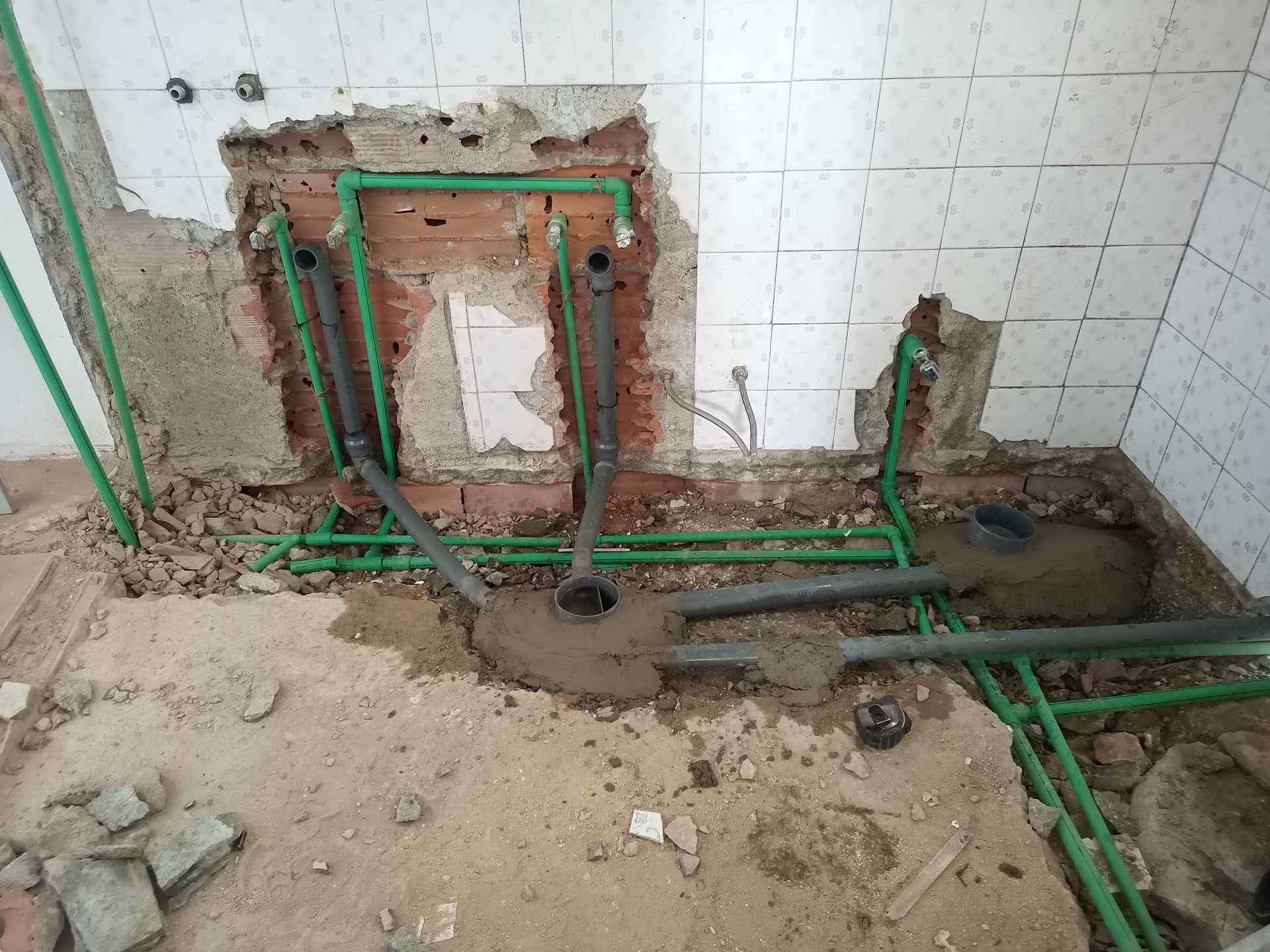 Reforma hidráulica em andamento: canos verdes e cinzas expostos na parede e no piso, com entulho.