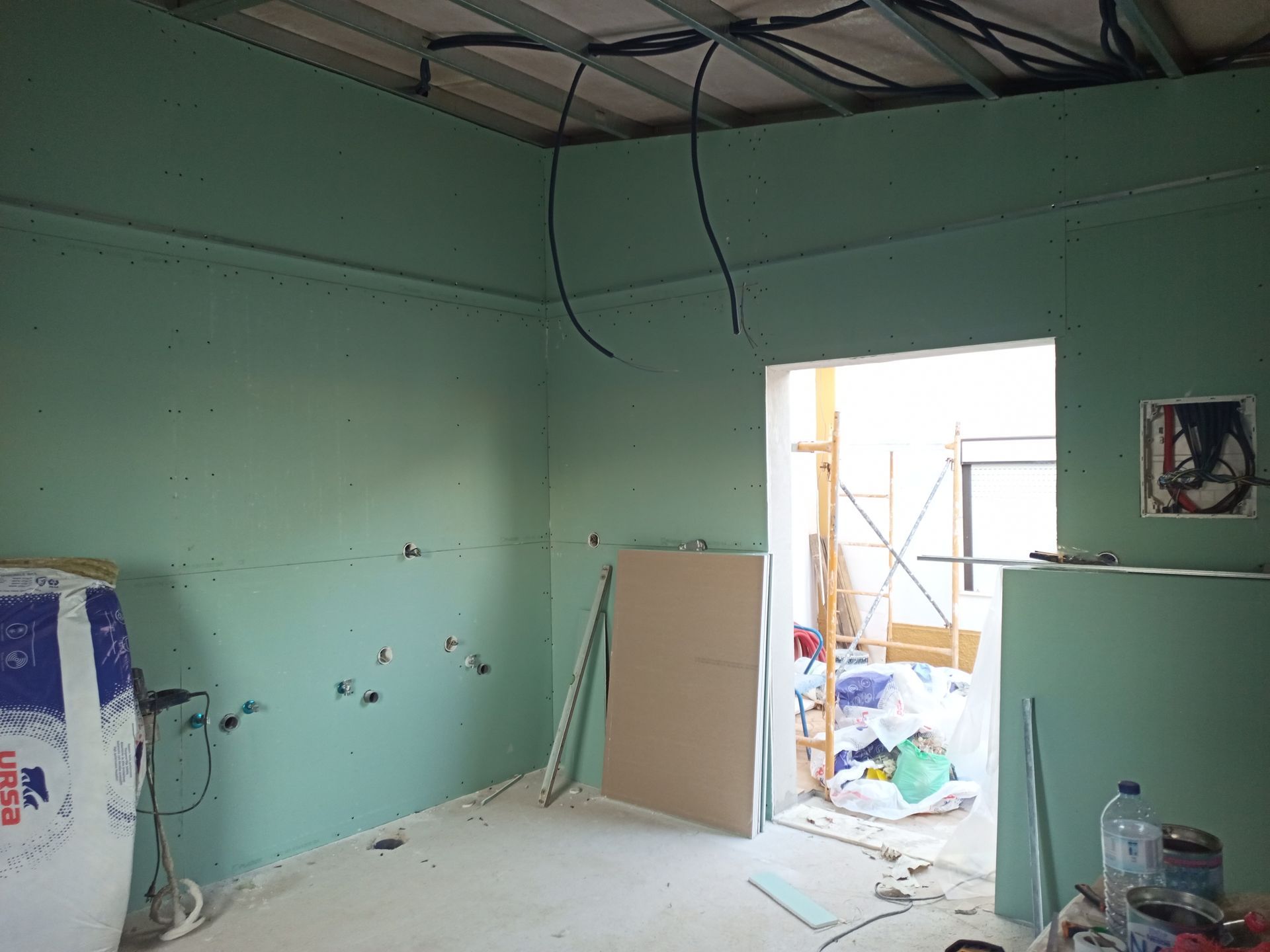 Sala em construção com paredes de gesso cartonado verde, fiação aparente e porta aberta.