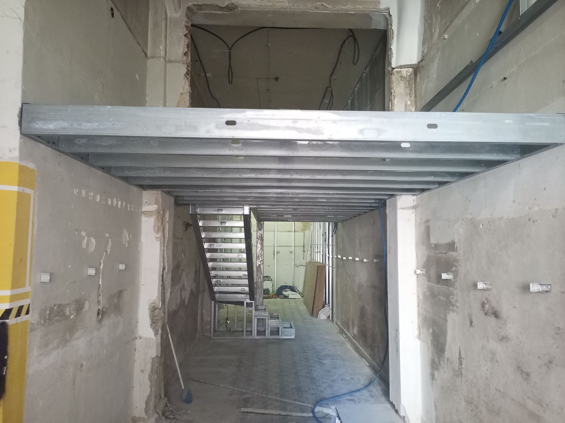 Construção interior: Estrutura de aço para um mezanino em um corredor, paredes de concreto inacabadas, escadas ao fundo.