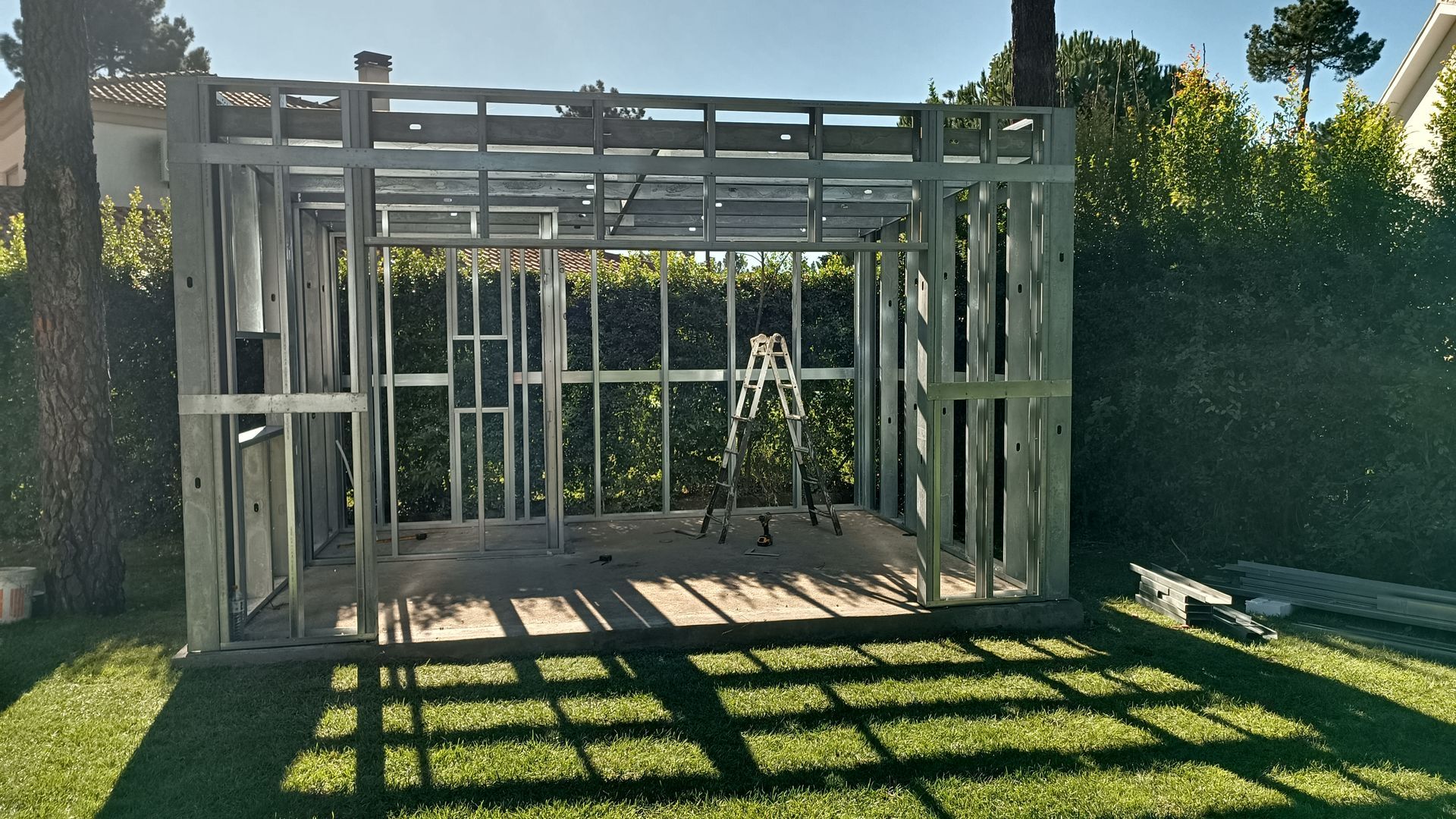 Estrutura metálica de um pequeno galpão em construção, situado em um gramado ensolarado.