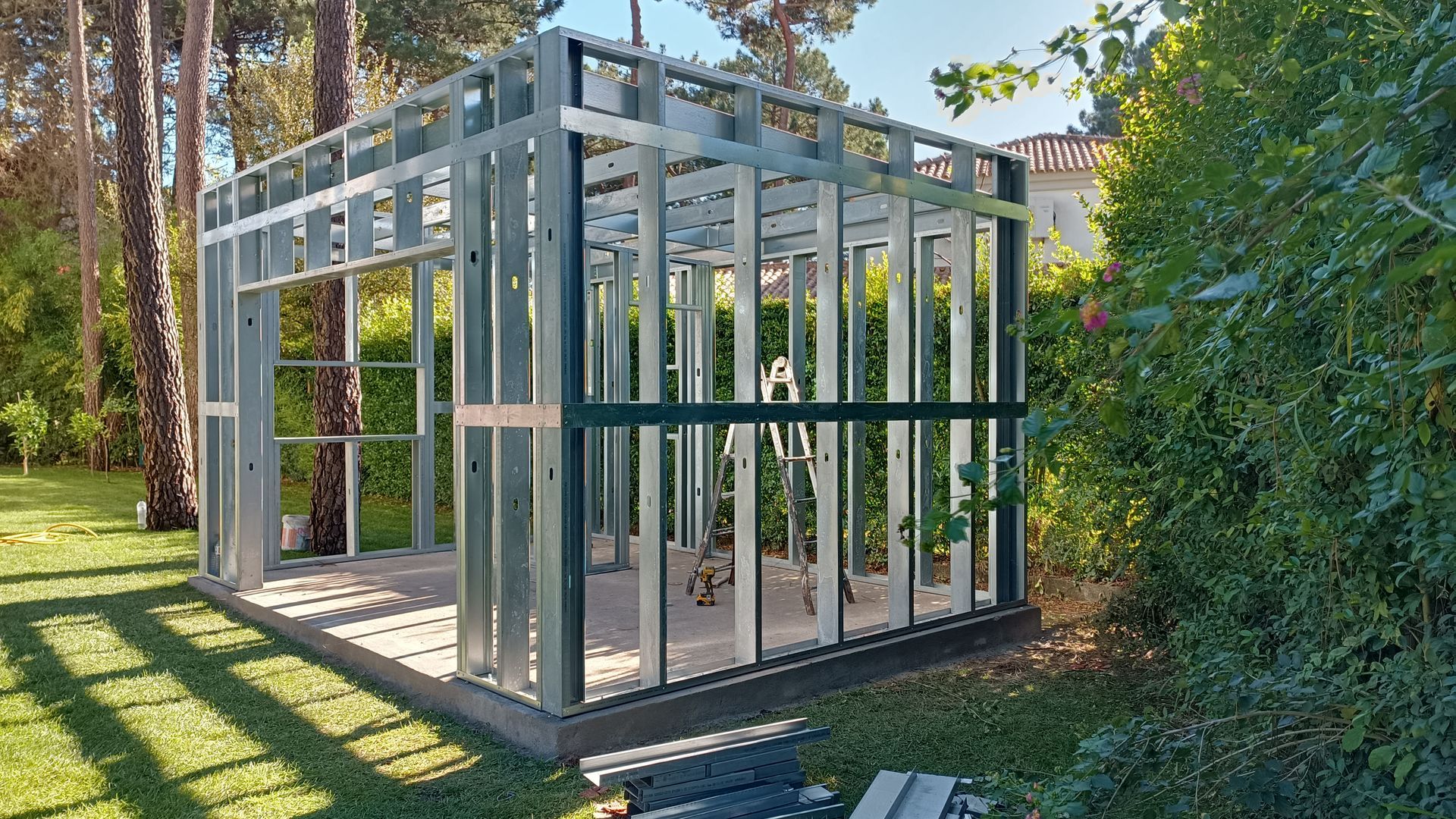 Estrutura de aço de um pequeno edifício em construção, em um pátio gramado, cercado por árvores.