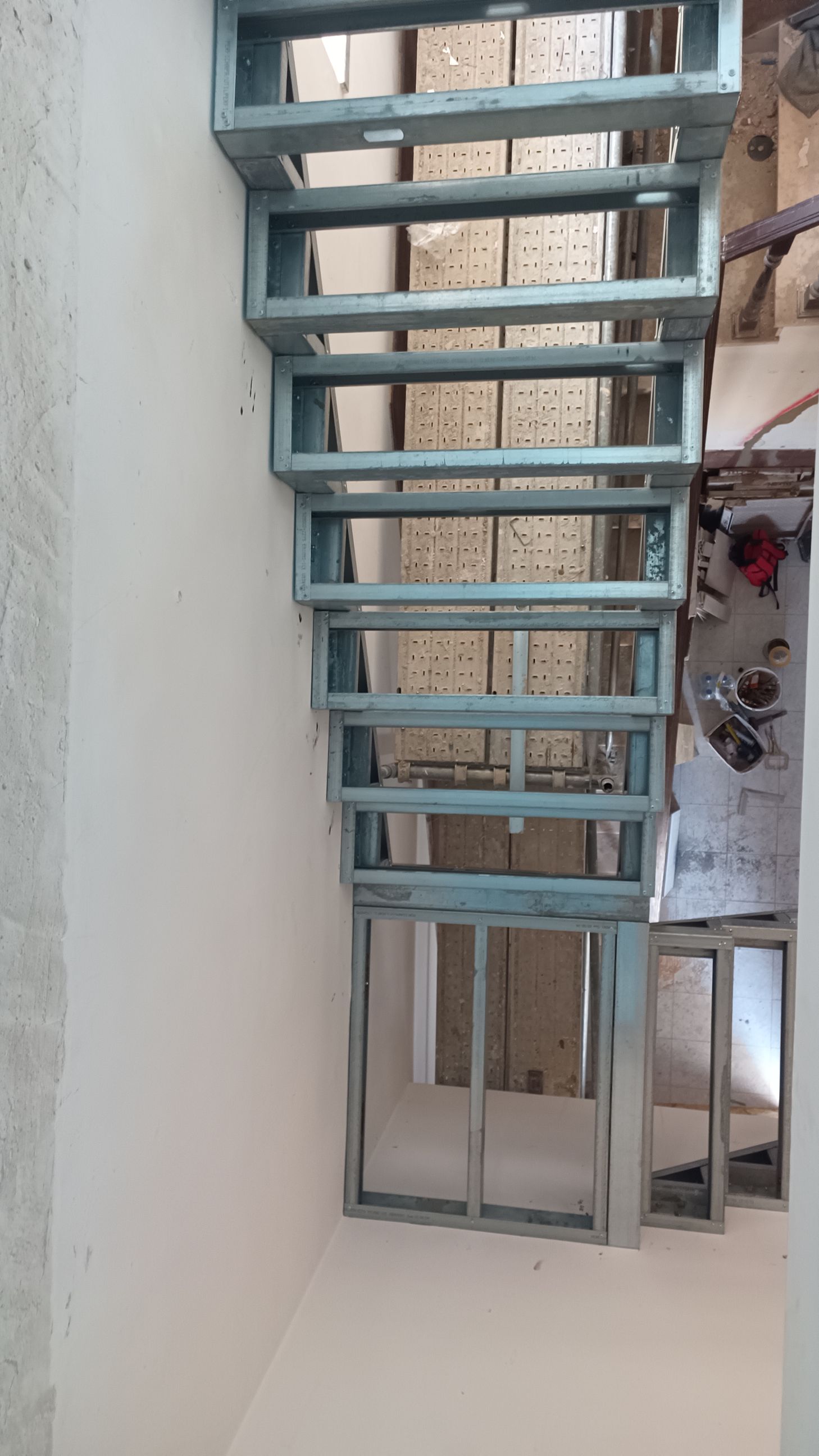 Estrutura metálica da escada, em tom azul-acinzentado, instalada contra uma parede branca. A escada leva a um nível inferior.