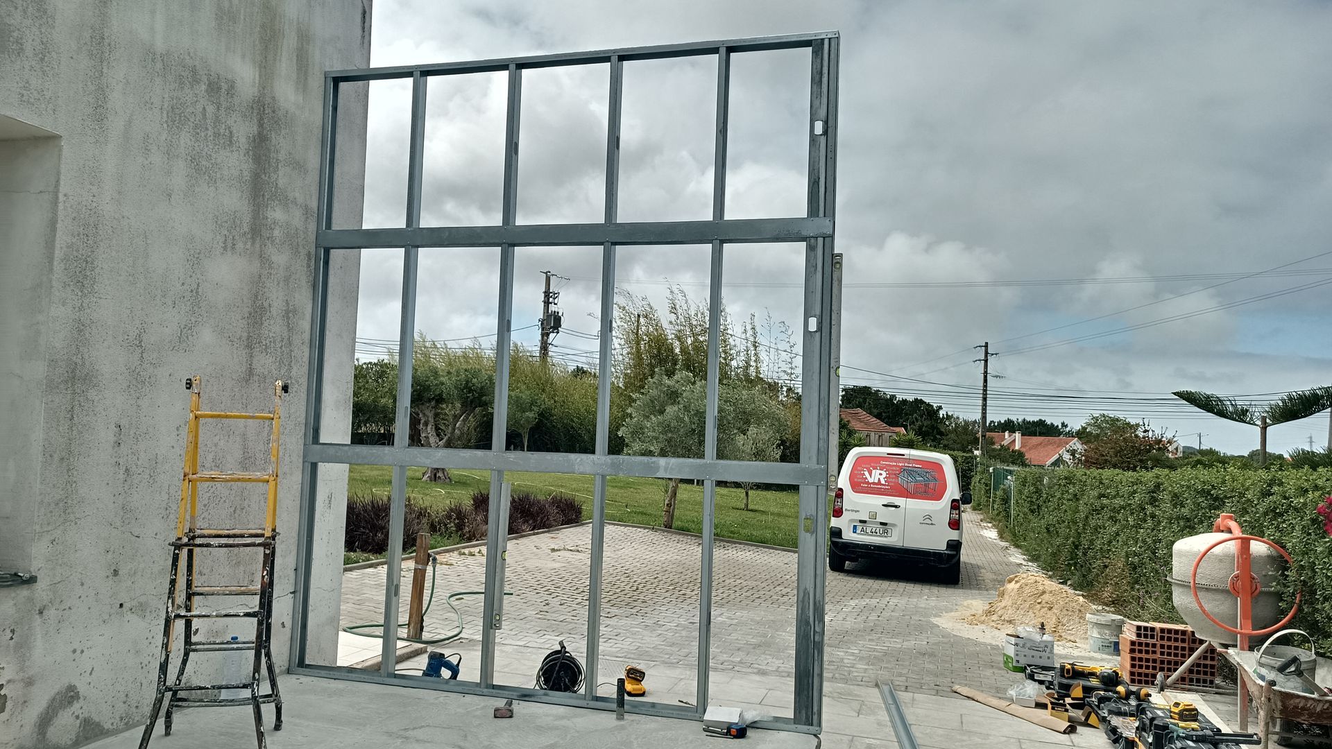 Estrutura metálica para uma grande janela ou porta, em construção perto de uma casa. Ferramentas de construção estão por perto.