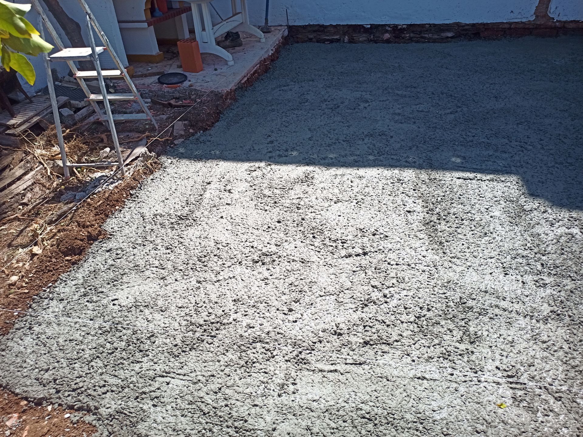 Pátio de concreto recém-construído, parcialmente coberto com um revestimento verde claro, adjacente a um terreno de terra e a um edifício.