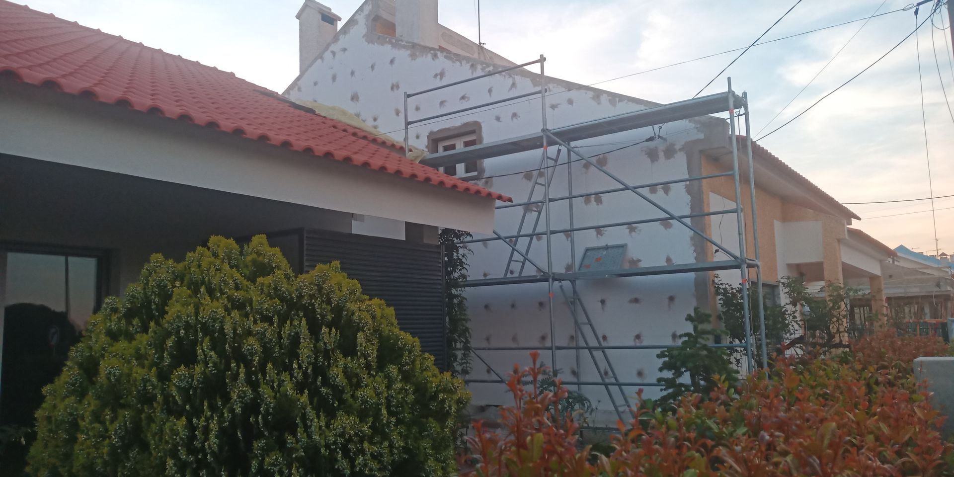 Edifício com andaimes, painéis isolantes e telhado de telha vermelha. Árvores e céu visíveis.