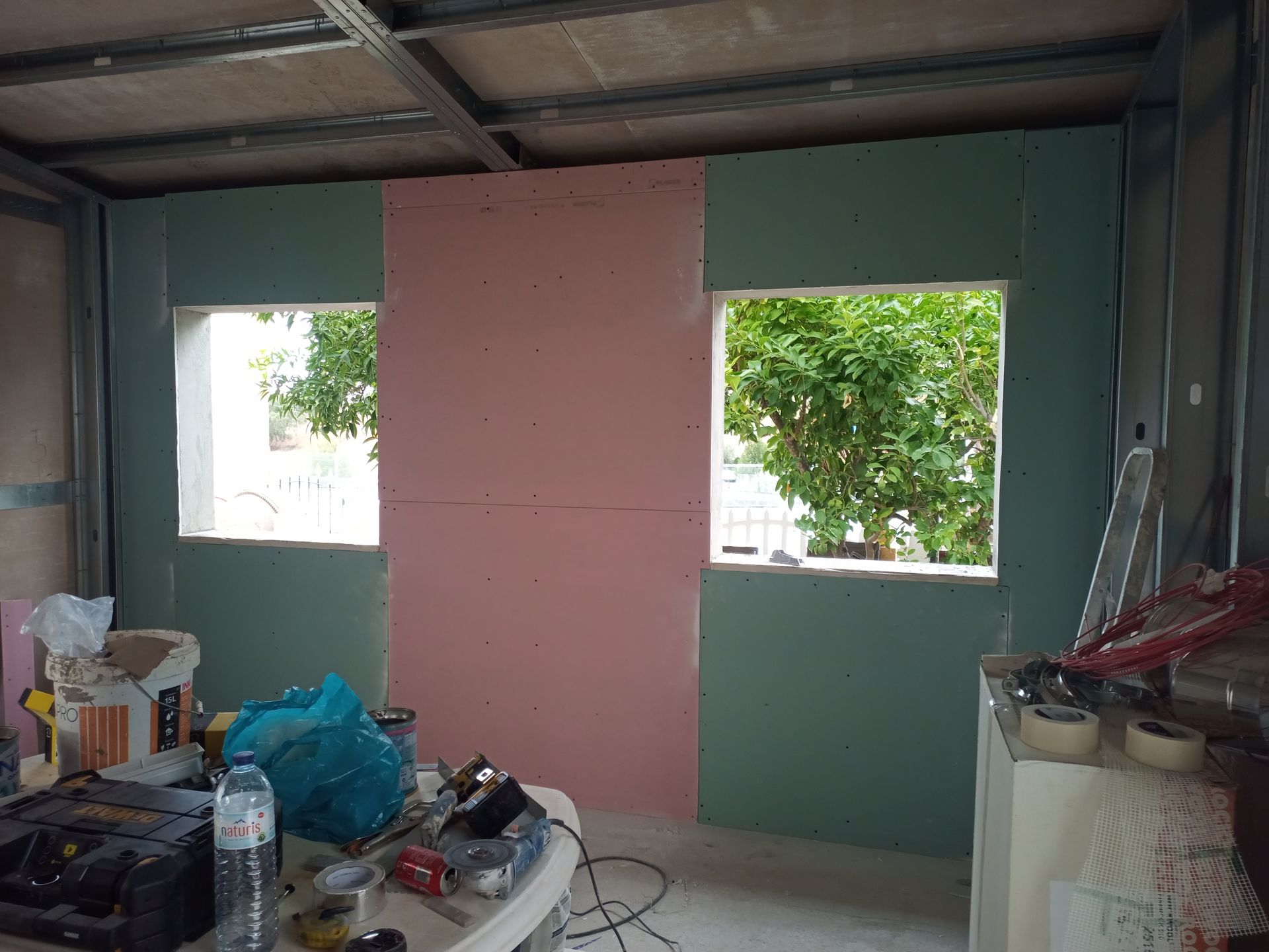 Cena de construção interna: Placas de gesso verdes e rosas sobre perfis metálicos, duas janelas, ferramentas e materiais sobre uma mesa.