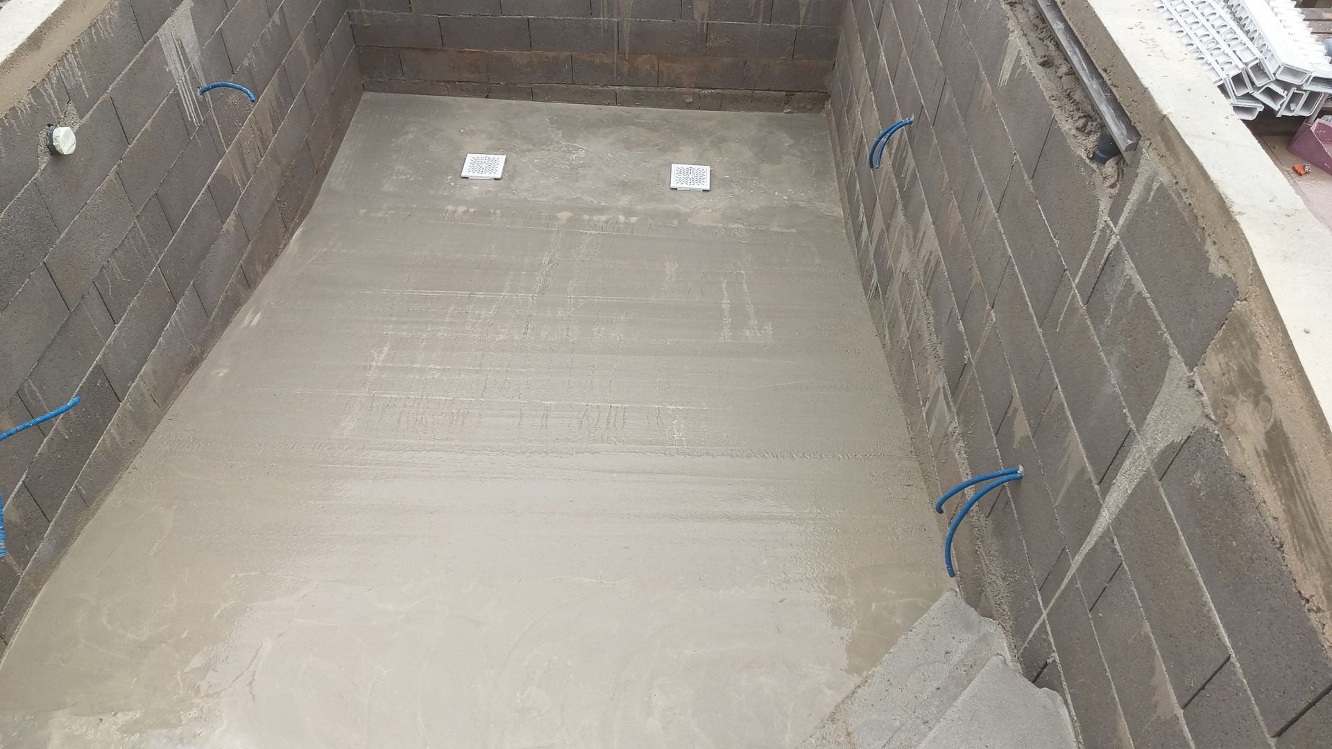 Vista interna de uma sala de blocos de concreto com piso de concreto. Fios azuis são visíveis nas paredes.