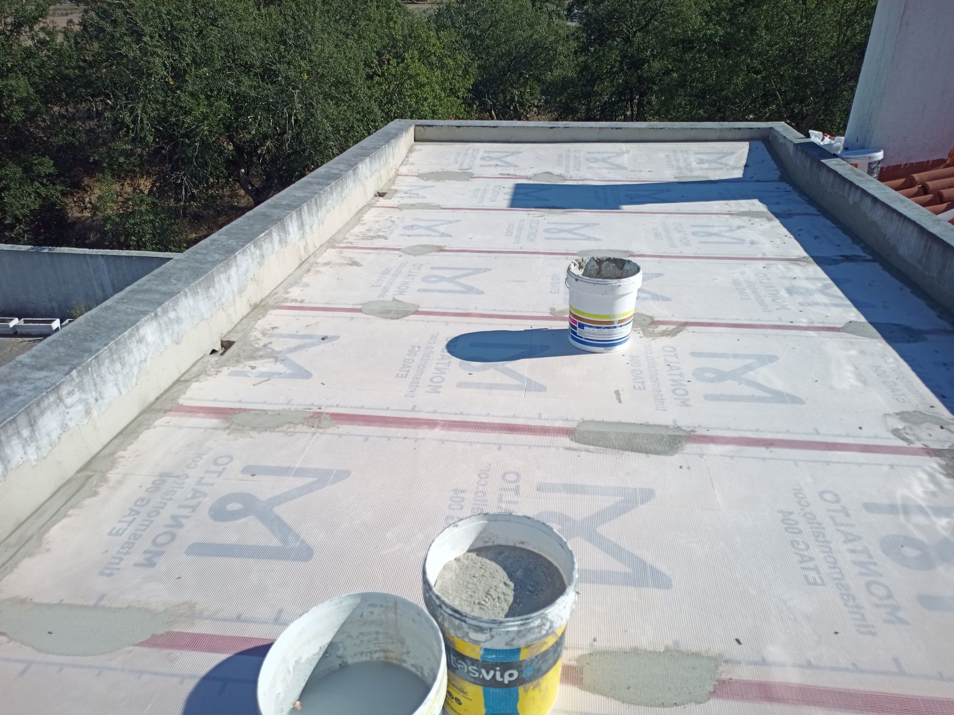 Telhado plano em construção; baldes de material, lonas protetoras brancas e uma borda de concreto.