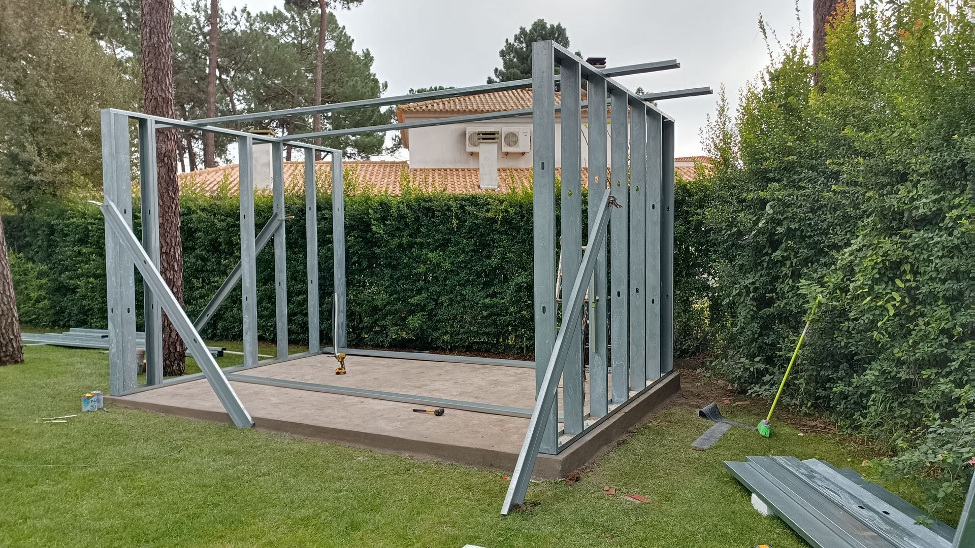 Estrutura metálica de um pequeno galpão em construção, situado em um gramado, com árvores ao redor e uma casa ao fundo.