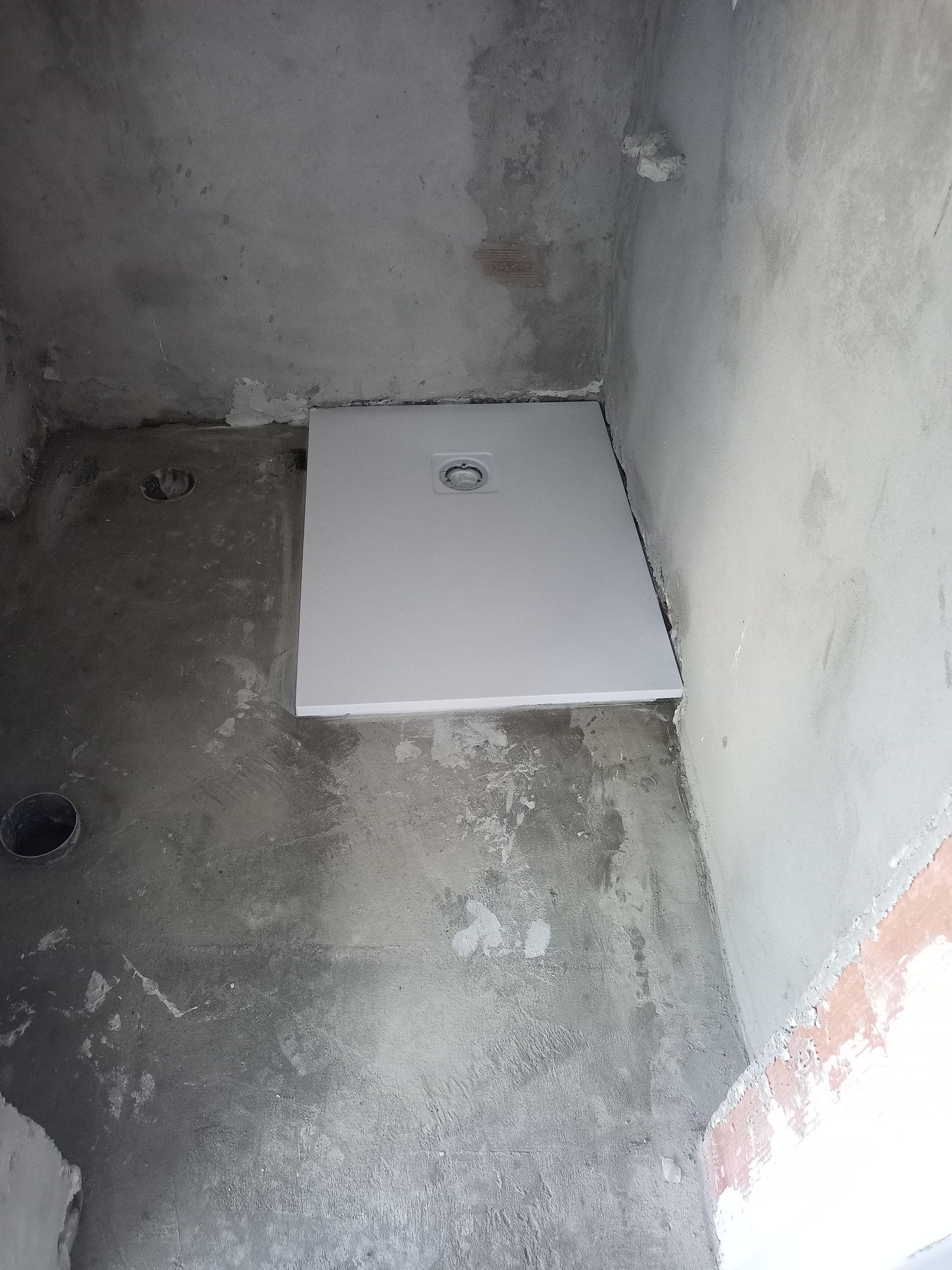 Um banheiro pequeno e inacabado com uma base de chuveiro branca instalada. Paredes e piso de concreto.