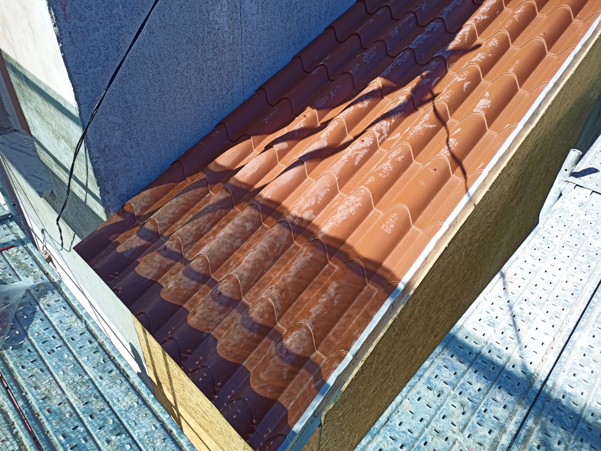 Telhado de telha marrom com juntas visíveis, luz solar e parte de um canteiro de obras.