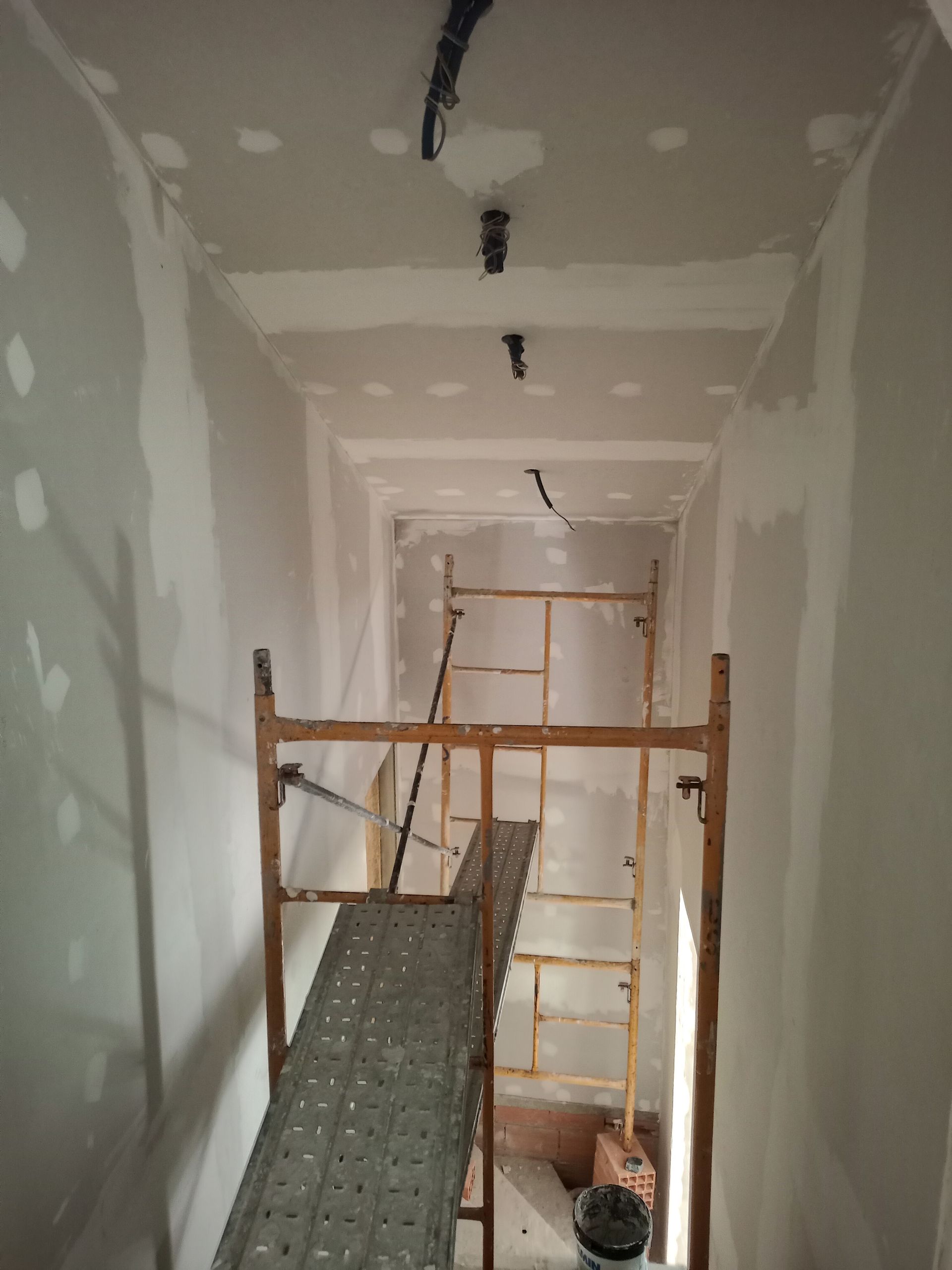Corredor em construção com andaimes. Paredes e teto com placas de gesso acartonado instaladas; iluminação visível no teto.