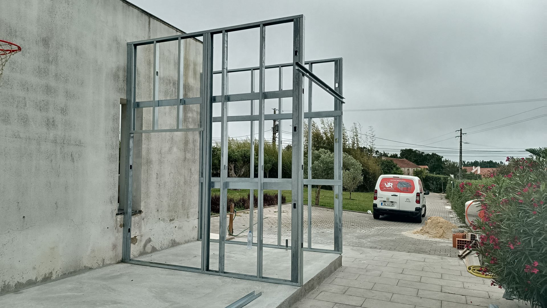 Estrutura metálica de um edifício em construção ao ar livre, ao lado de uma parede existente, céu nublado.