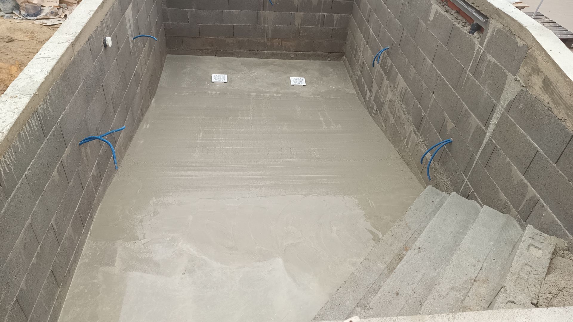 Construção de uma piscina de concreto. Paredes e degraus de blocos de concreto cinza. Tubos azuis salientes.