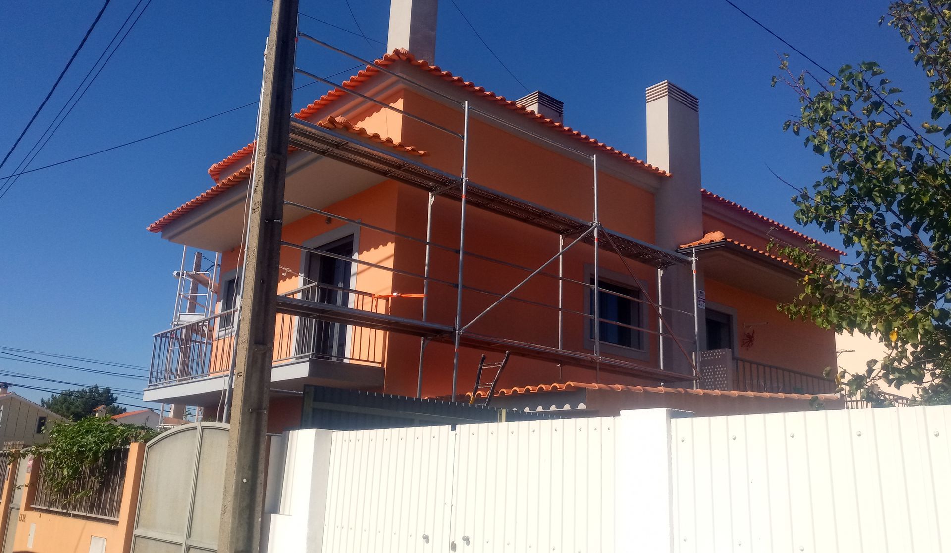 Casa laranja de dois andares em construção, com andaimes e telhado de telha vermelha, contra um céu azul claro.