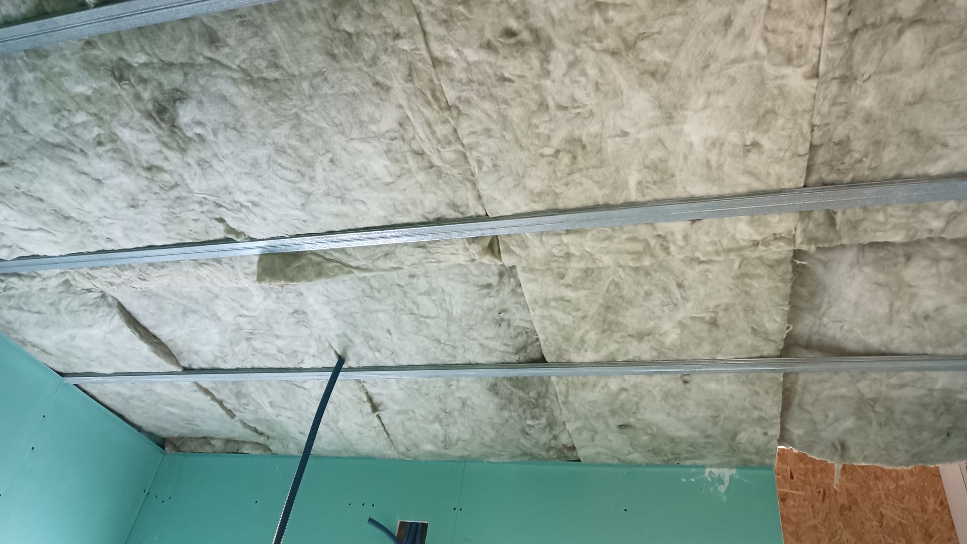 Isolamento instalado entre as estruturas metálicas do teto. Parede verde visível à esquerda, painéis de madeira à direita.