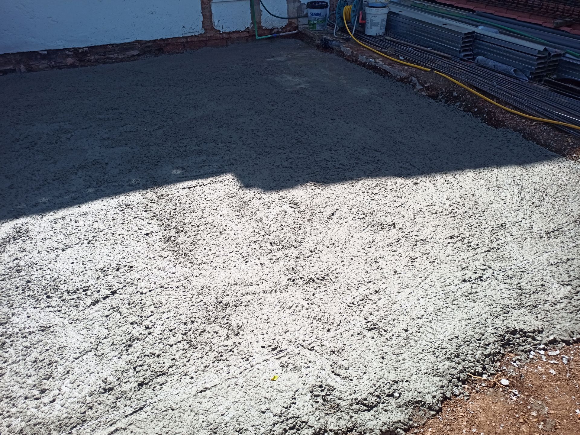 Superfície de concreto recém-lançado. Uma seção está na sombra, revelando sua textura. Materiais de construção estão ao fundo.