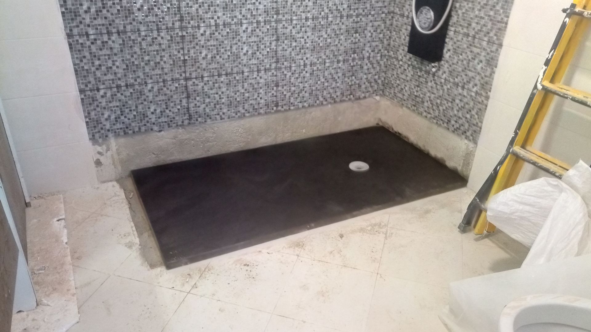 Box de chuveiro em construção, com base preta, parede revestida com azulejos cinza e piso branco.