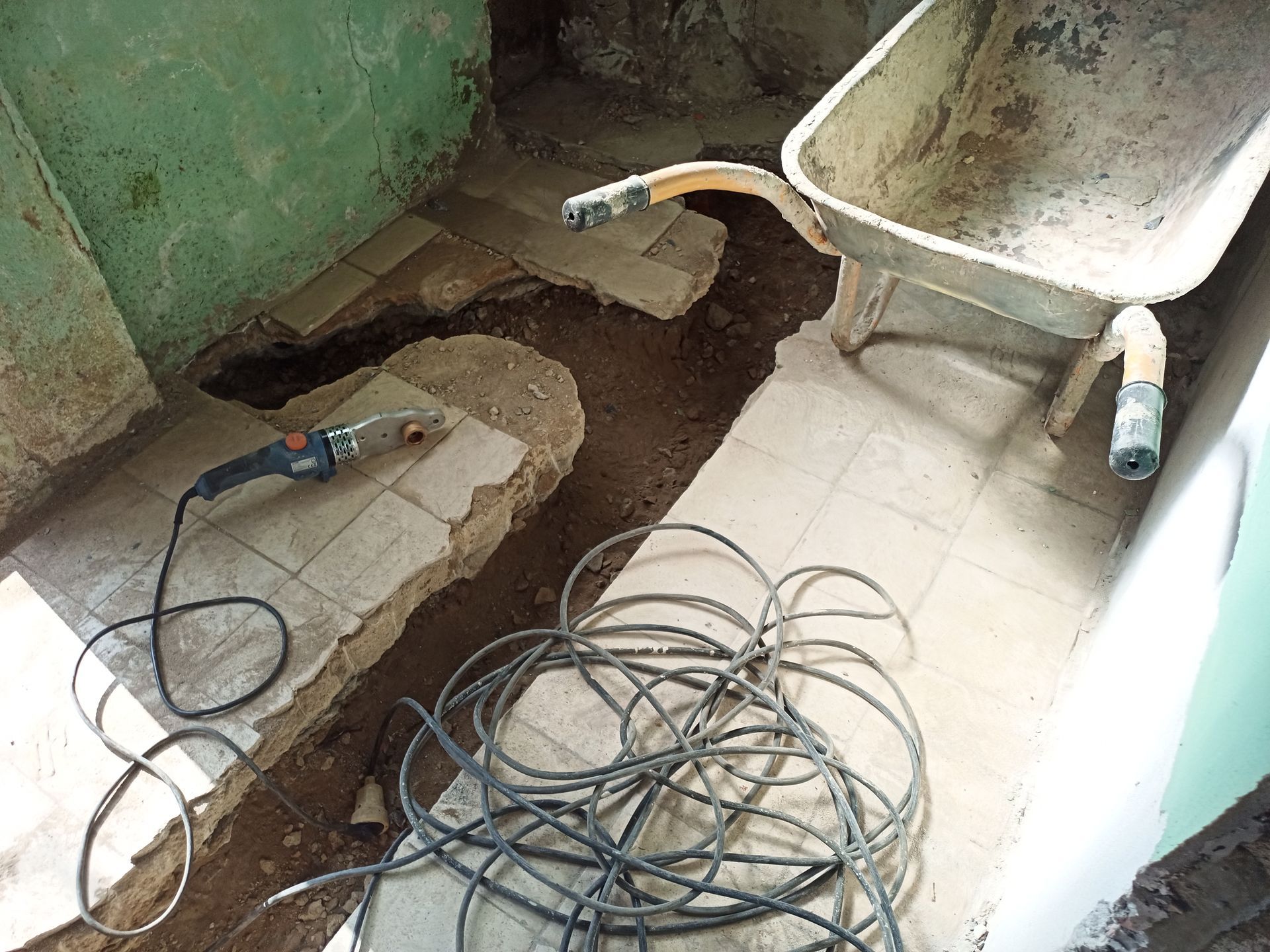 Piso de azulejos com uma vala, um carrinho de mão, uma ferramenta elétrica e fios emaranhados, em uma sala com paredes verdes.