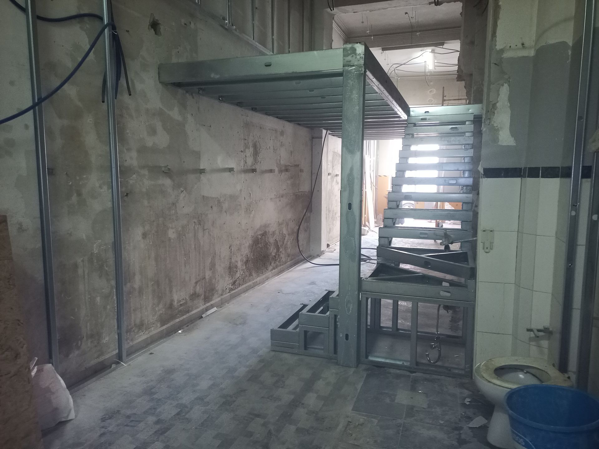 Escada e plataforma metálicas em um cômodo inacabado, com vigas expostas e um banheiro.