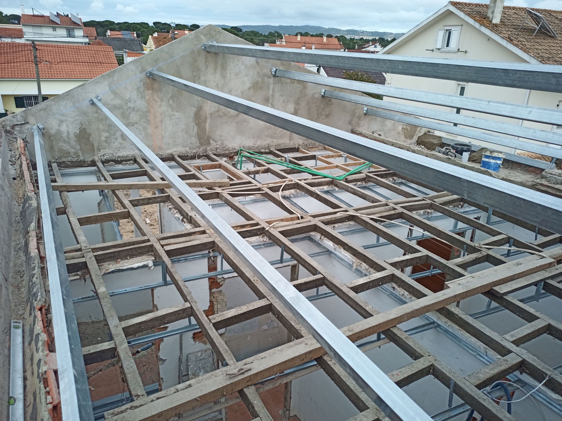 Canteiro de obras: estrutura do telhado com vigas metálicas e suportes de madeira, exposta ao céu, dia nublado.