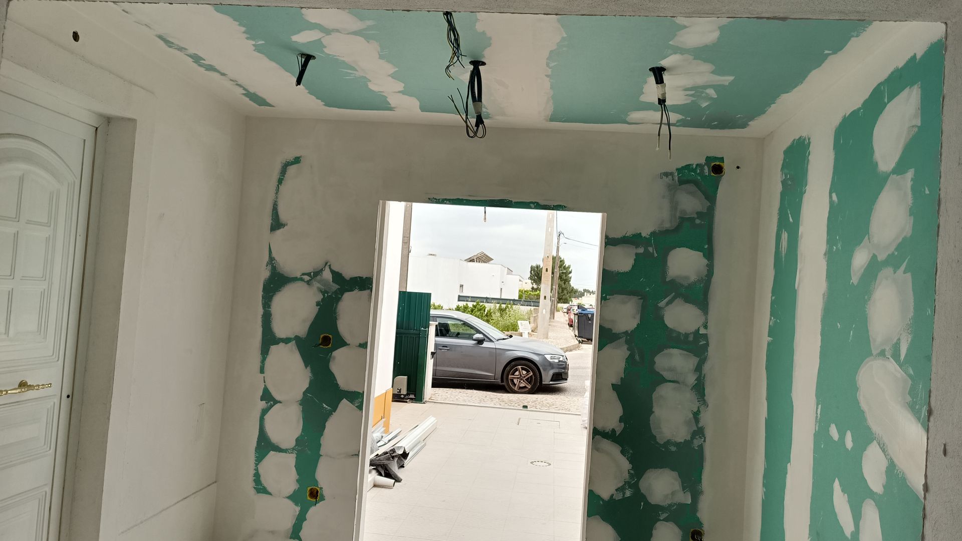Porta interna e teto em construção, com drywall e fiação aparentes. Um carro é visível do lado de fora.