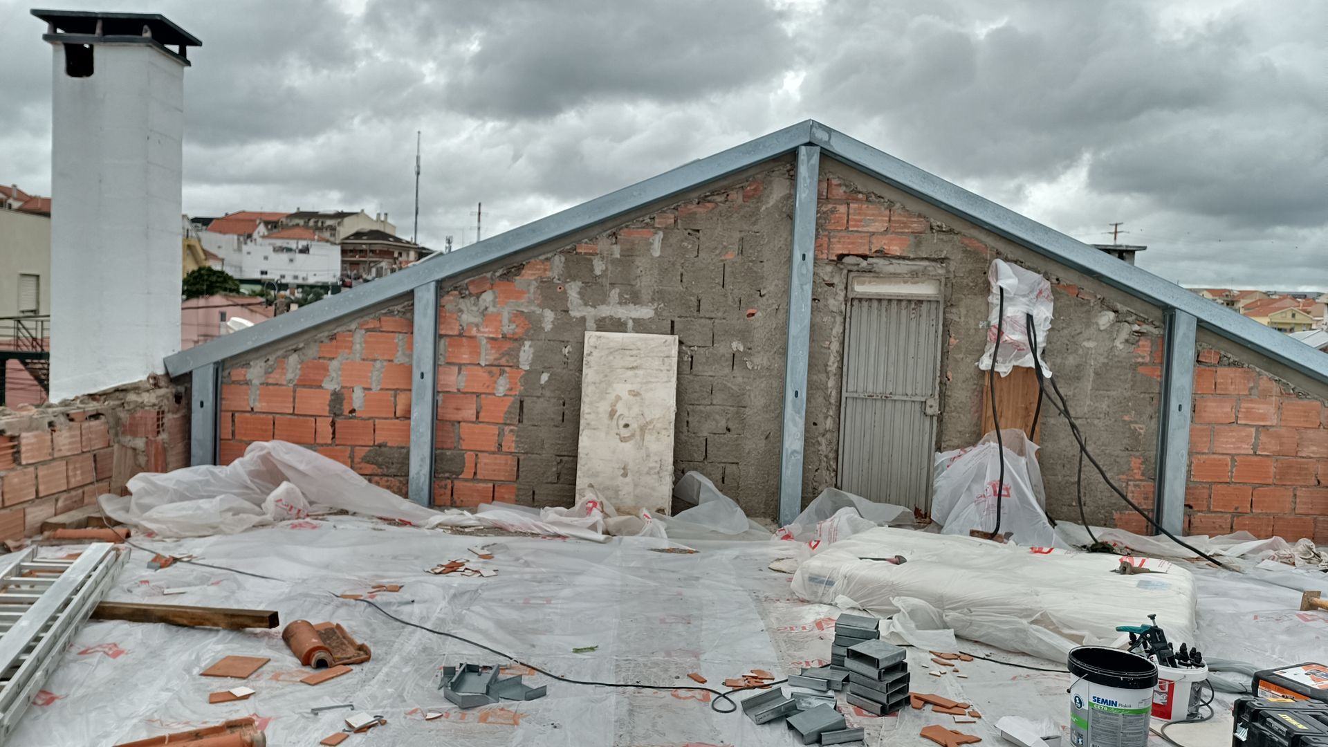 Construção no telhado com parede de tijolos, chaminé e céu nublado. Coberta com lona protetora e materiais de construção.