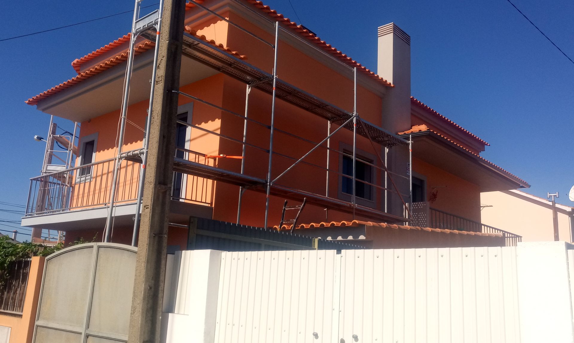 Casa laranja de dois andares com andaimes, sob um céu azul claro, atrás de uma cerca branca.