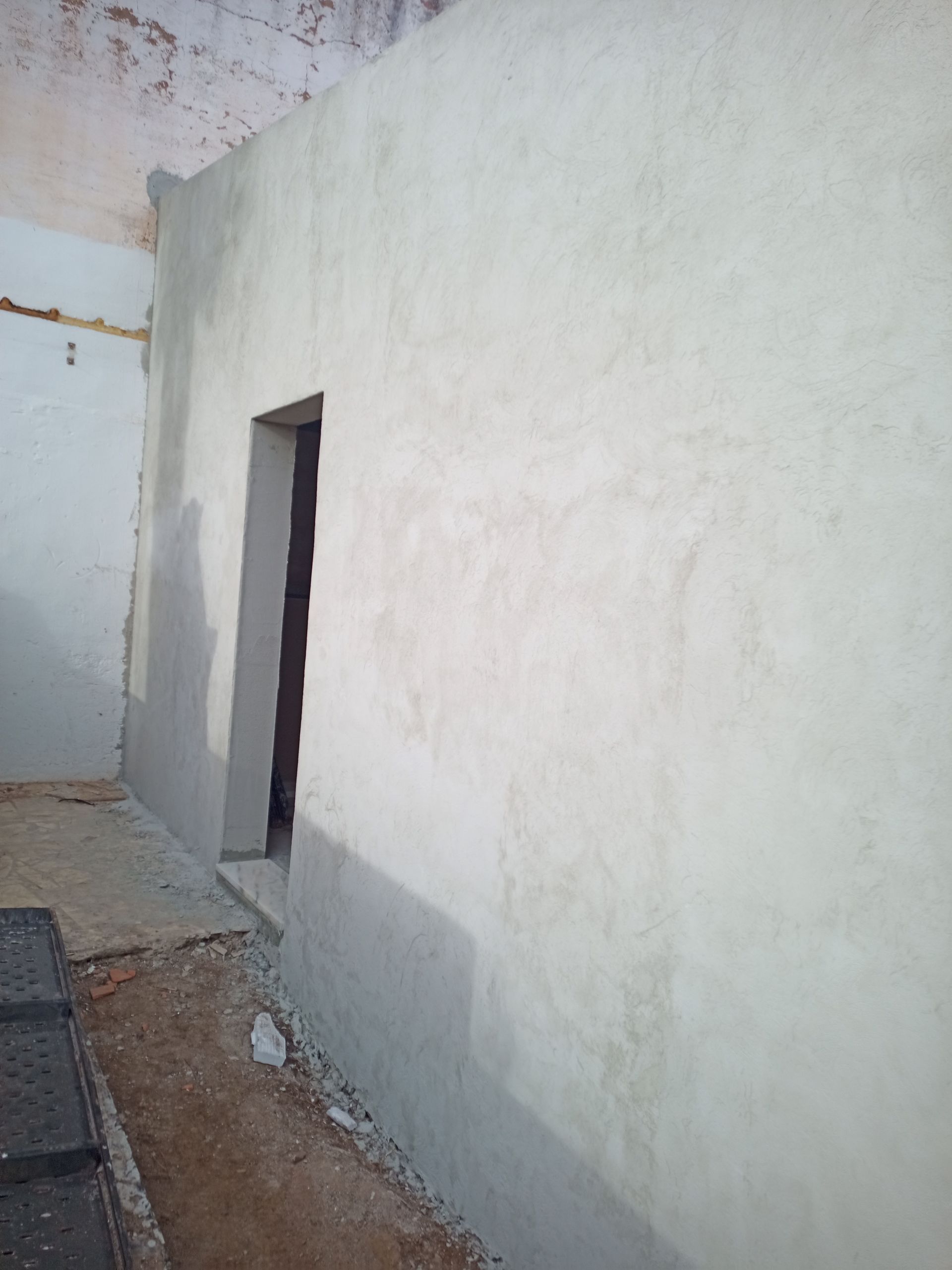 Quarto interior com paredes inacabadas e uma porta. Paredes bege, piso de concreto e luz natural.