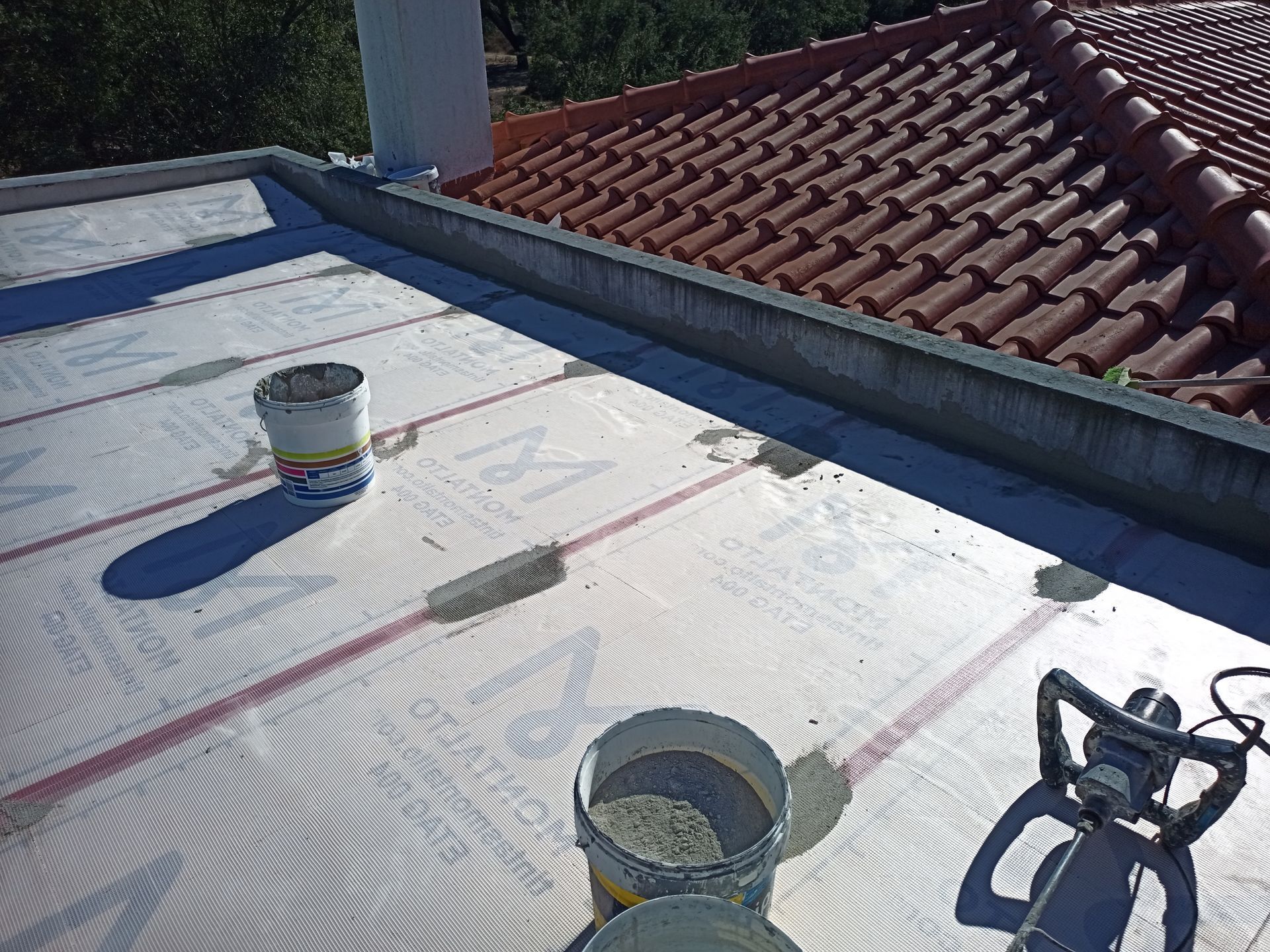 Construção de telhado plano: Baldes, selante e uma ferramenta elétrica estão sobre uma membrana branca, ao lado de um telhado de telhas.