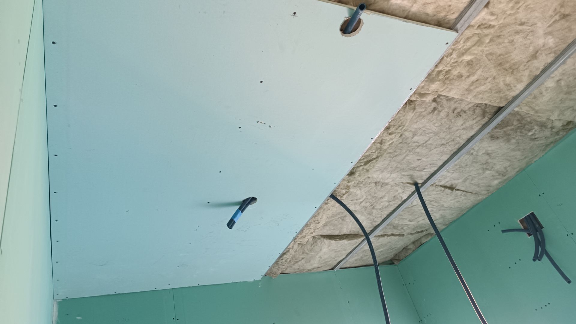 O teto está parcialmente coberto com gesso acartonado azul, revelando o isolamento e a estrutura metálica. Fios pendem do teto.