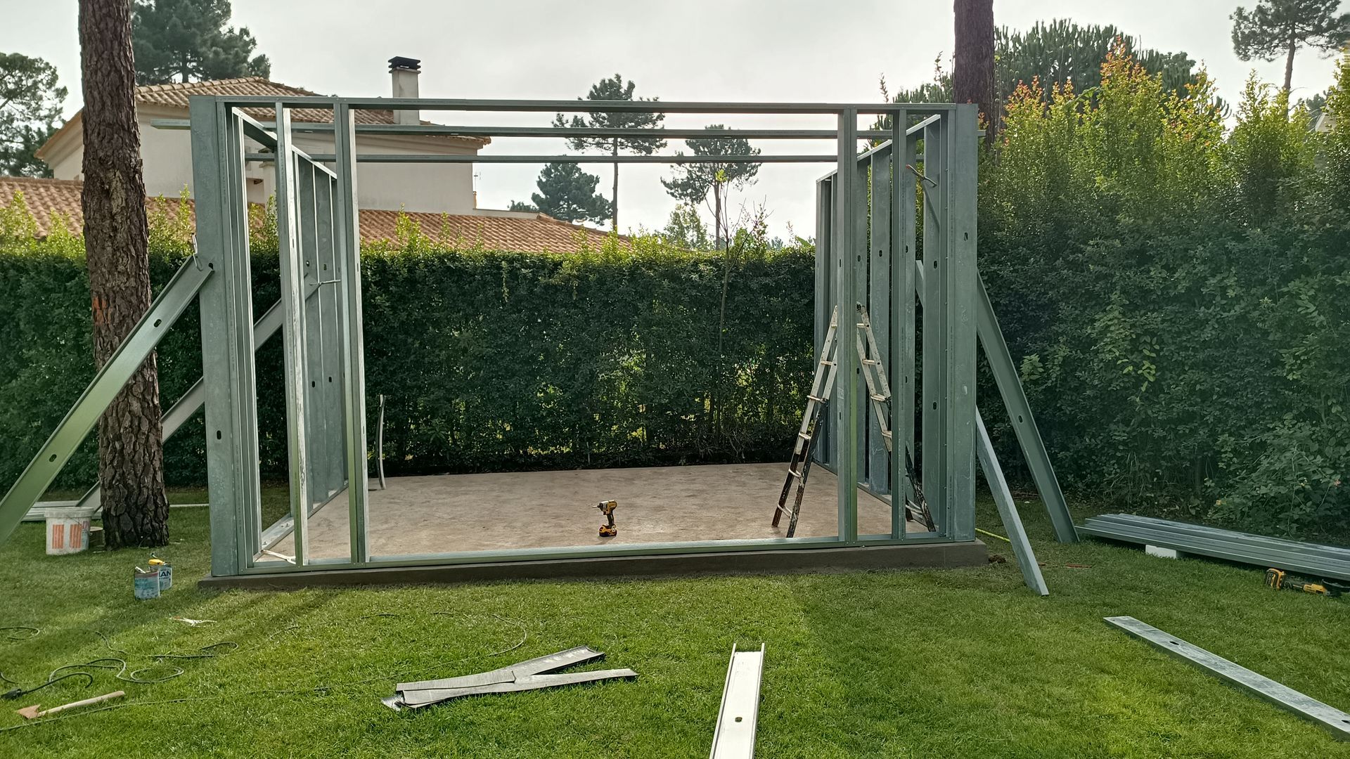 Galpão com estrutura metálica em construção em um quintal gramado, cercado por árvores e folhagens.