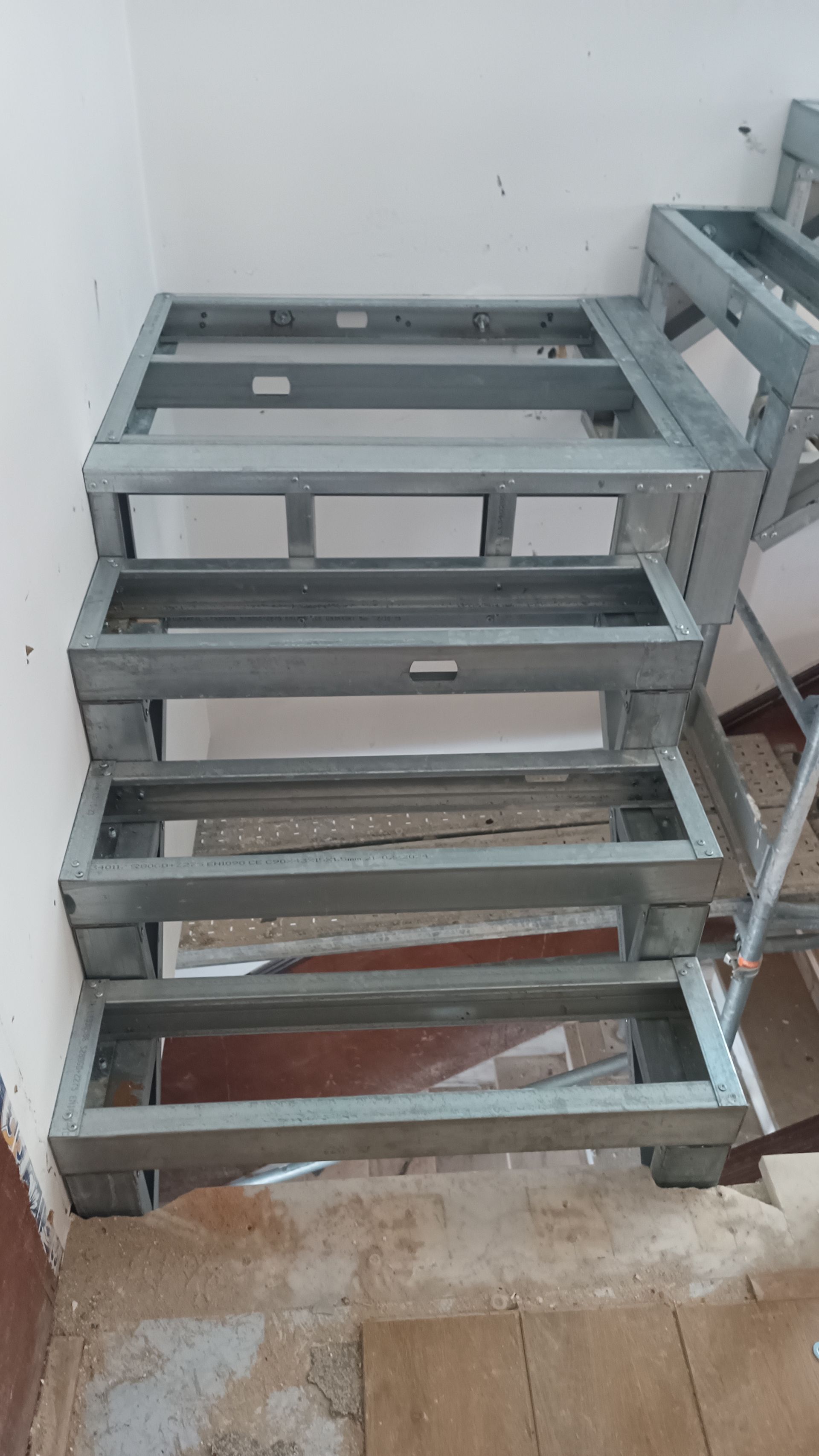 Estrutura metálica de escada em construção em uma sala com parede branca e piso de concreto.