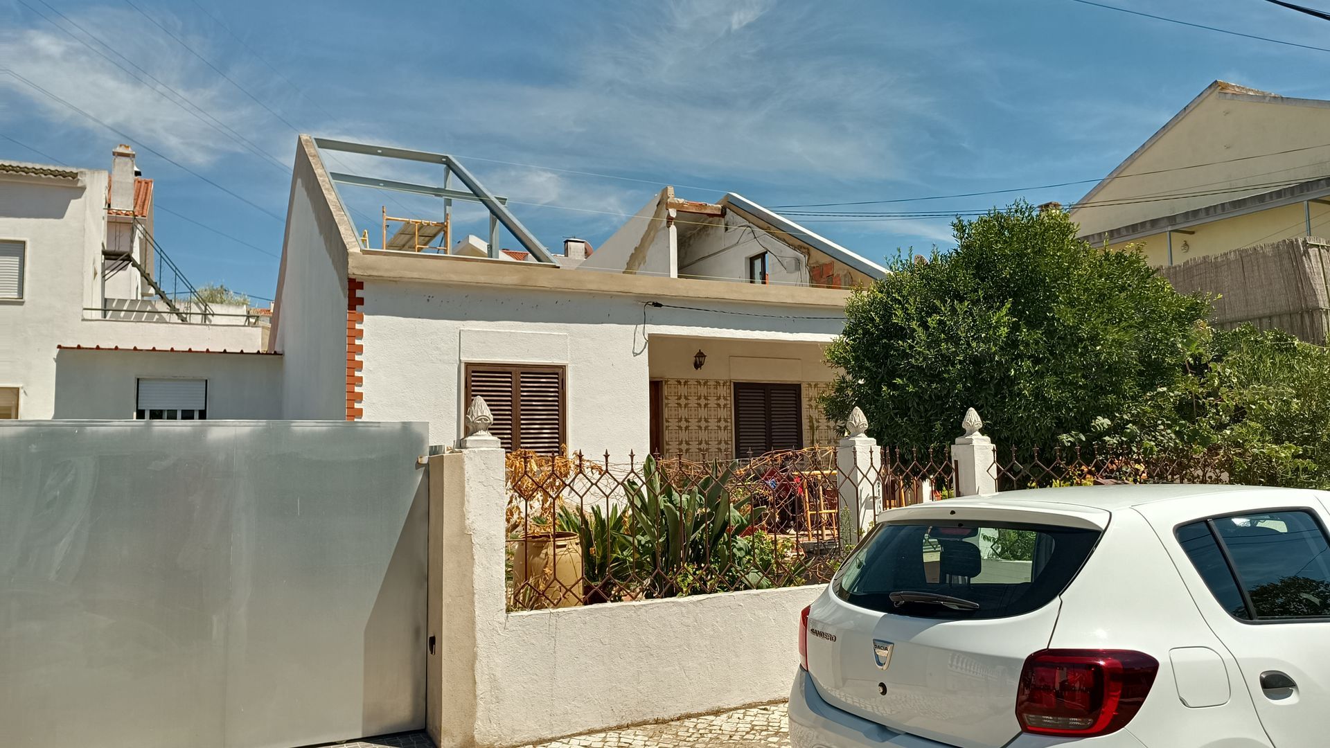 Casa branca com telhado parcialmente danificado, flores e um carro estacionado na frente em um dia ensolarado.