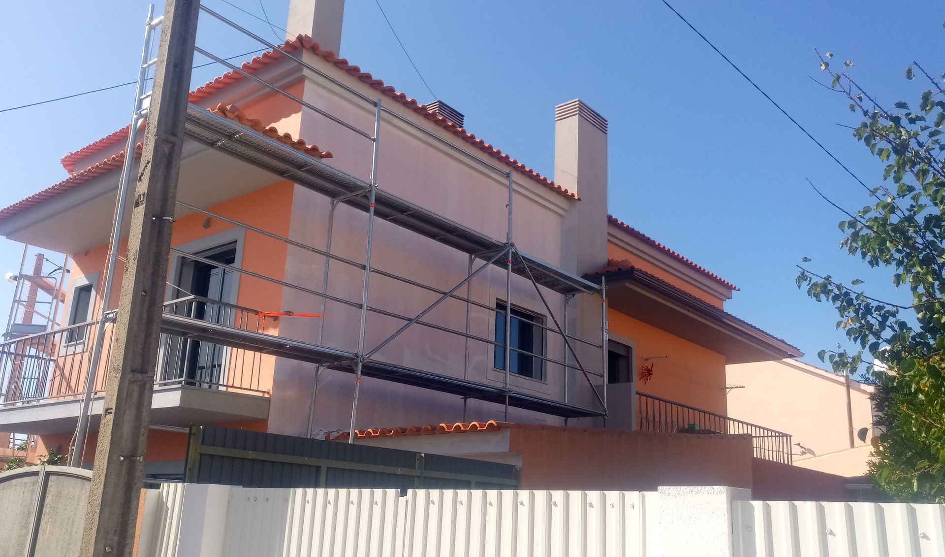 Prédio de dois andares em construção com andaimes. Exterior em estuque laranja, telhado e chaminé vermelhos. Dia ensolarado.