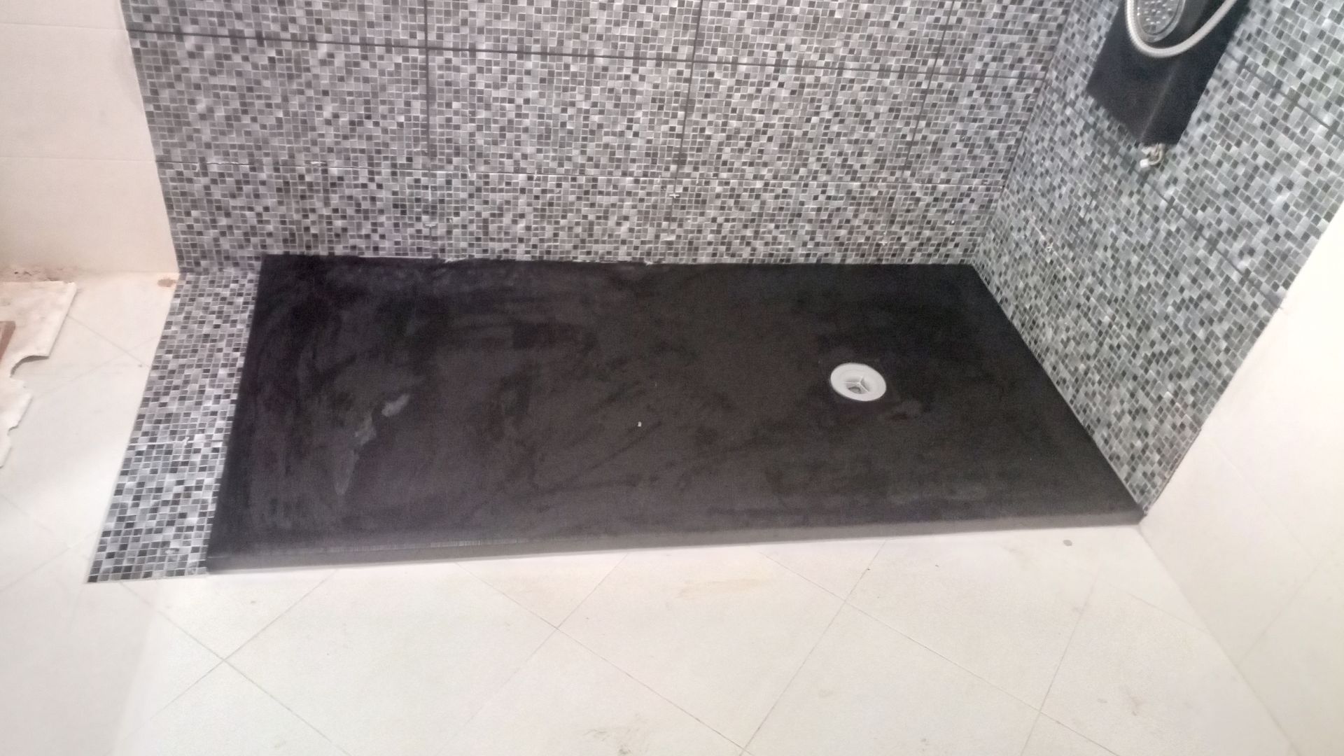 Base de chuveiro preta com ralo central, paredes revestidas com azulejos de mosaico cinza, piso branco.