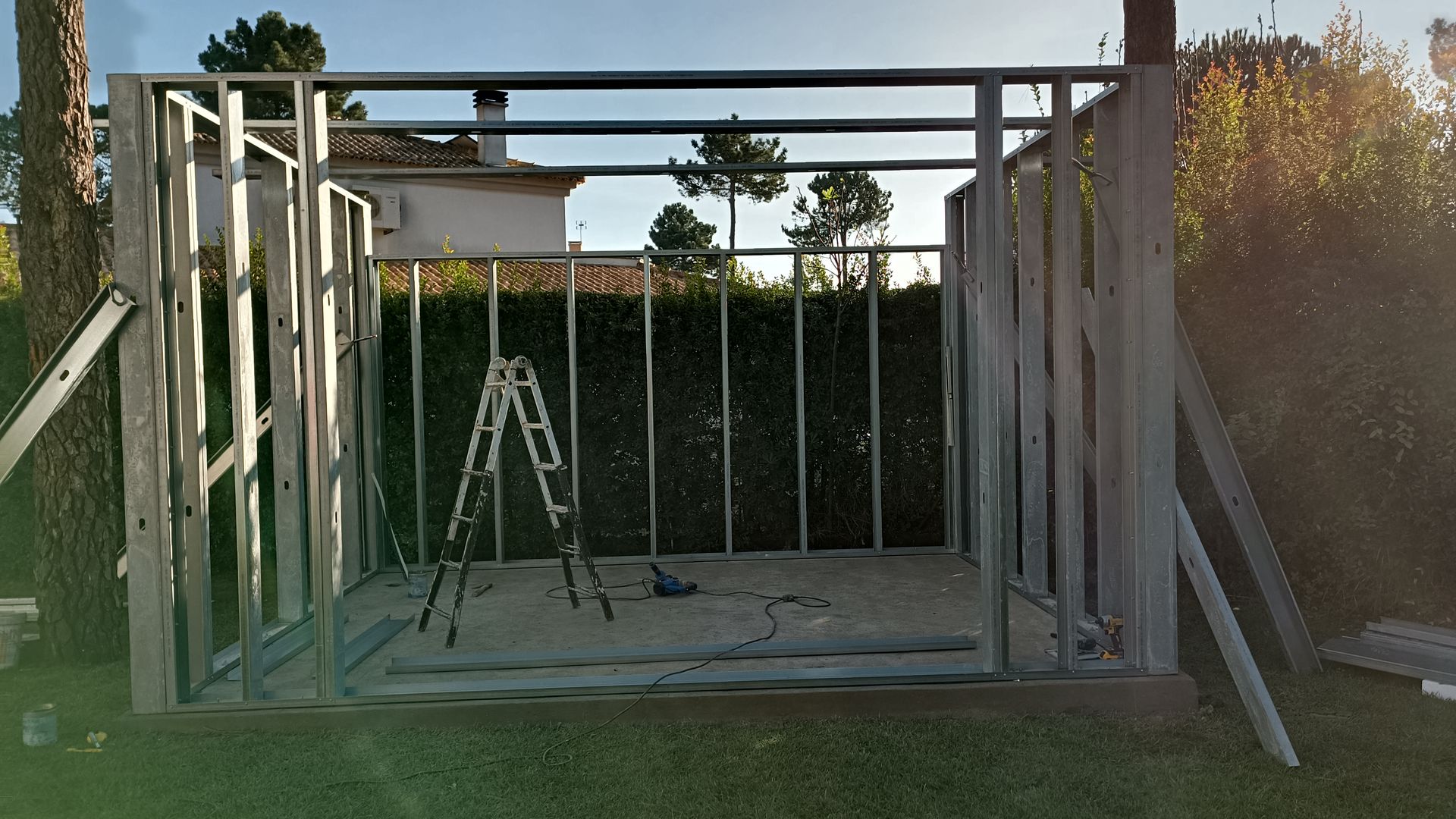 Estrutura metálica de um galpão em construção em um quintal gramado, com uma escada dentro.