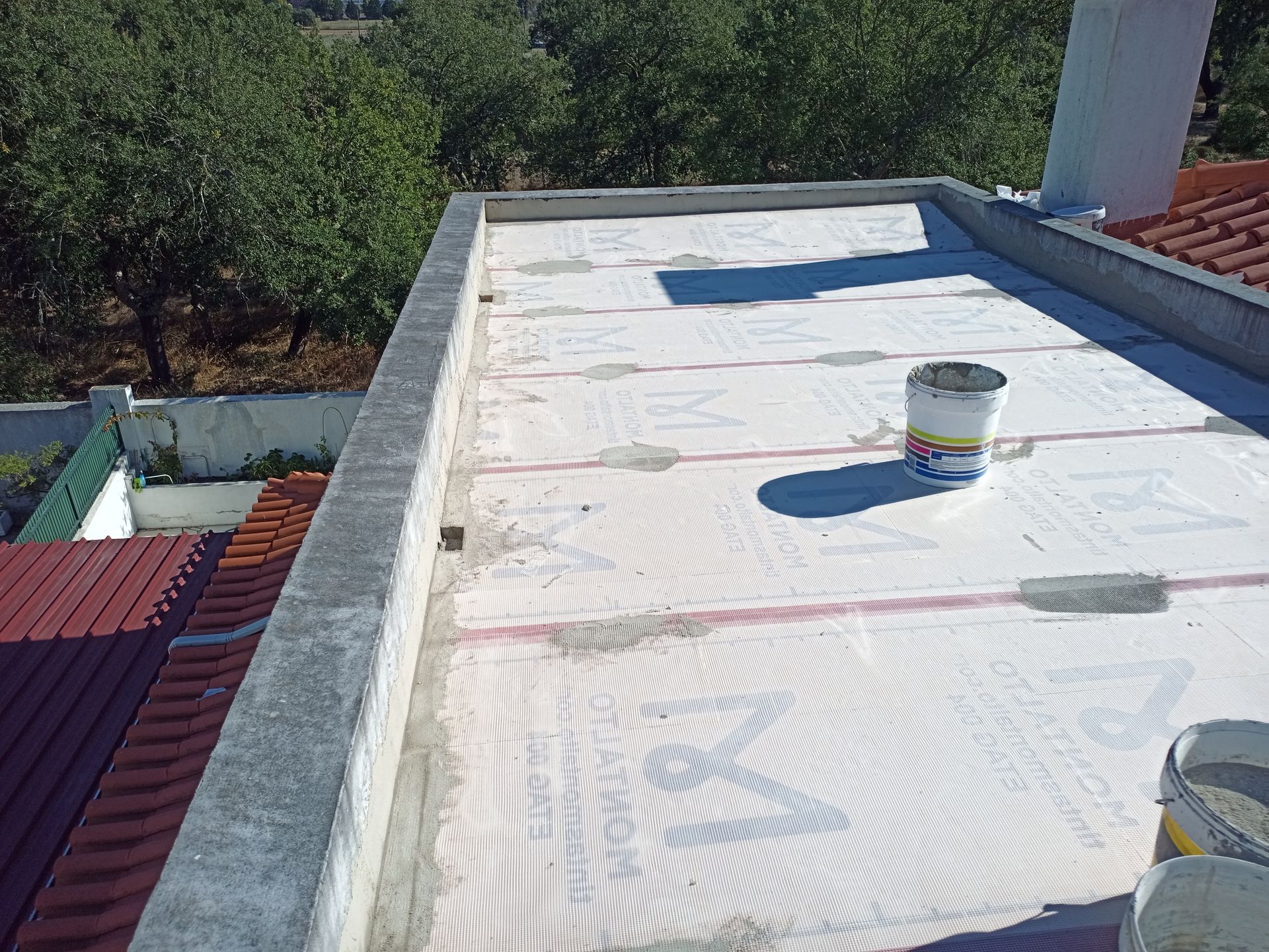 Telhado plano com tela branca e um balde de tinta. Árvores ao fundo.